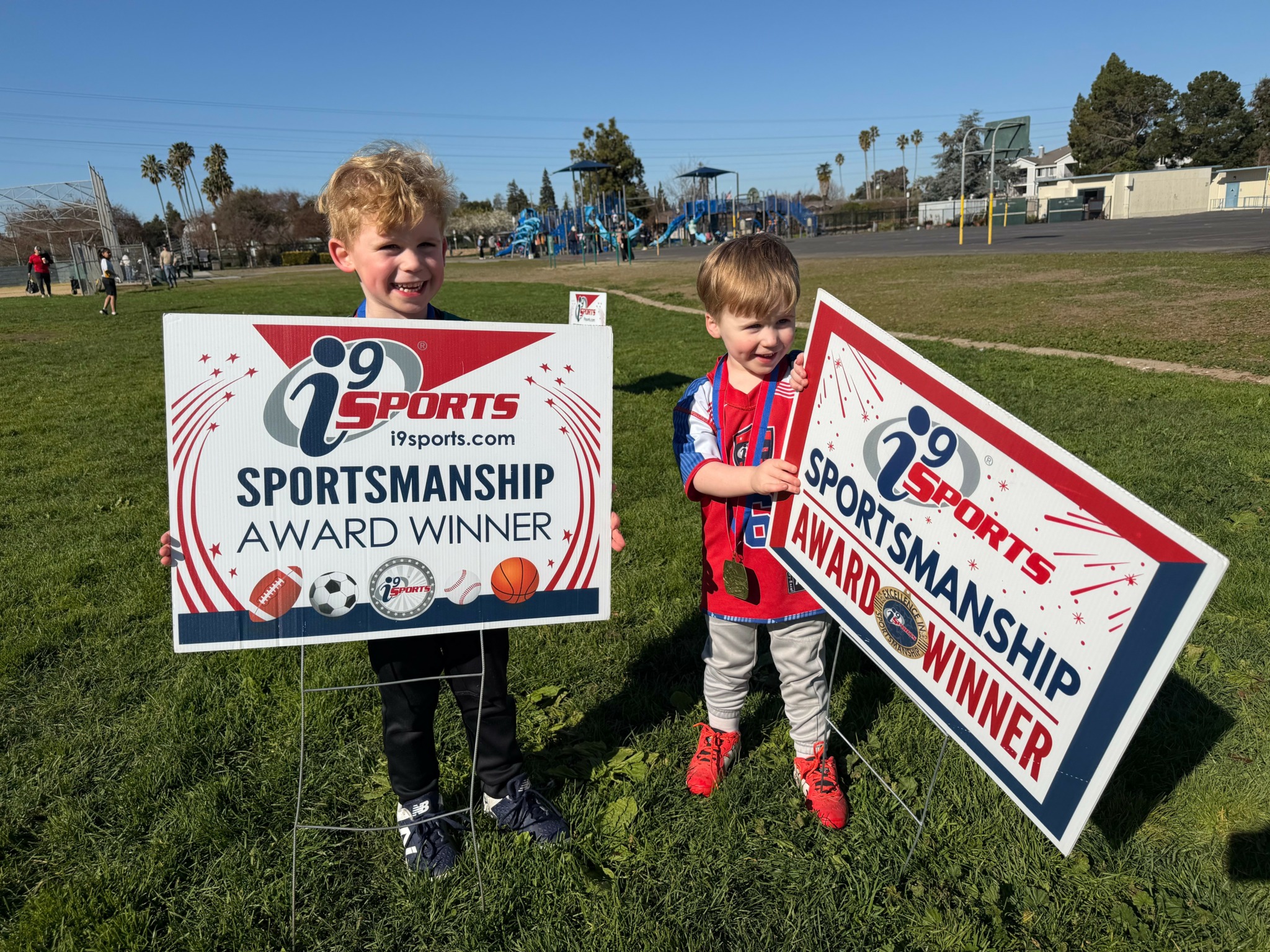 Two children holding signs that say 'Sportsmanship Award Winner' outdoors on a grassy field with a playground in the background.