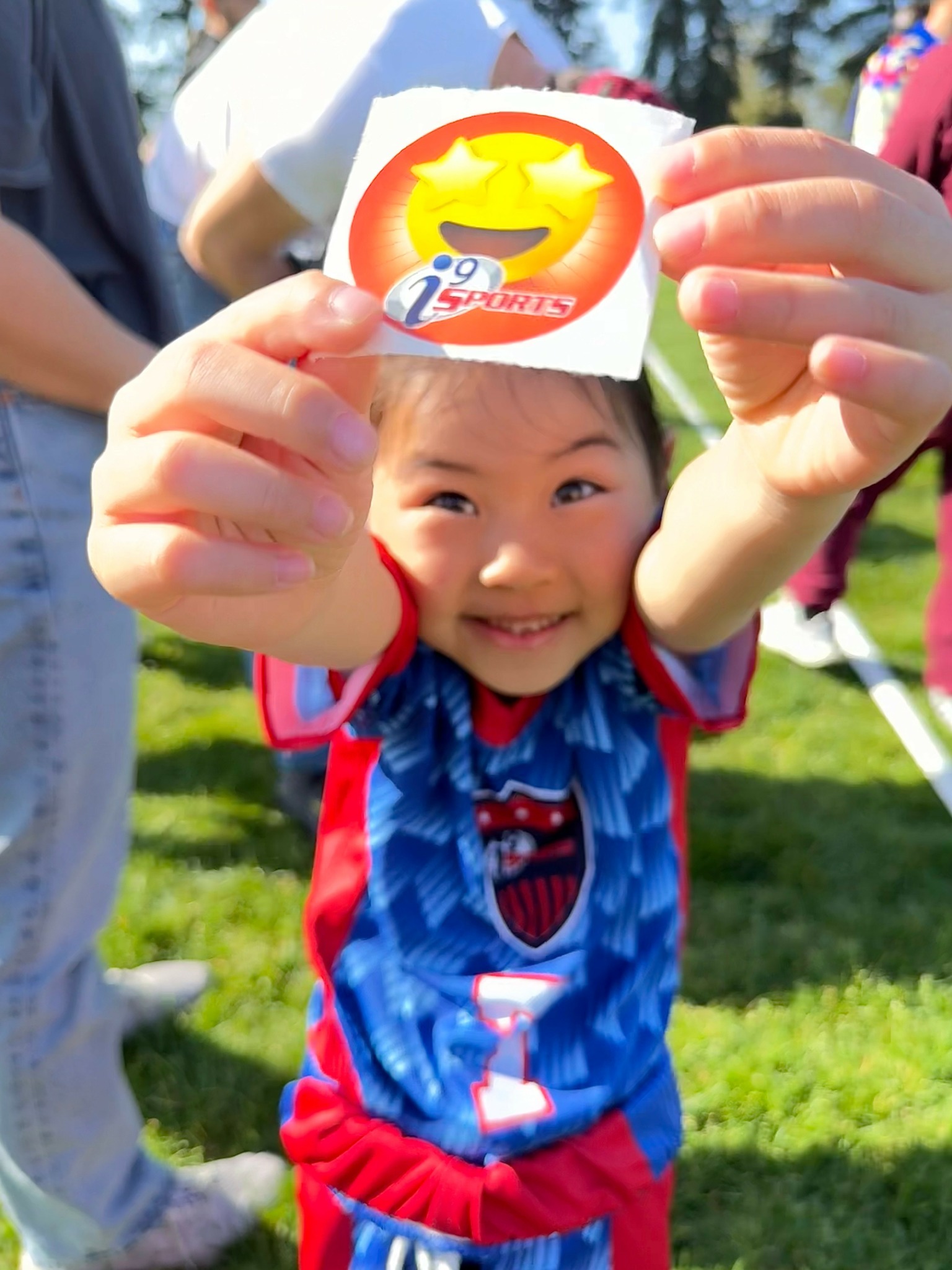 Child holding a sticker with a yellow star, smiling outdoors on grass, surrounded by people, wearing a blue and red sports outfit.