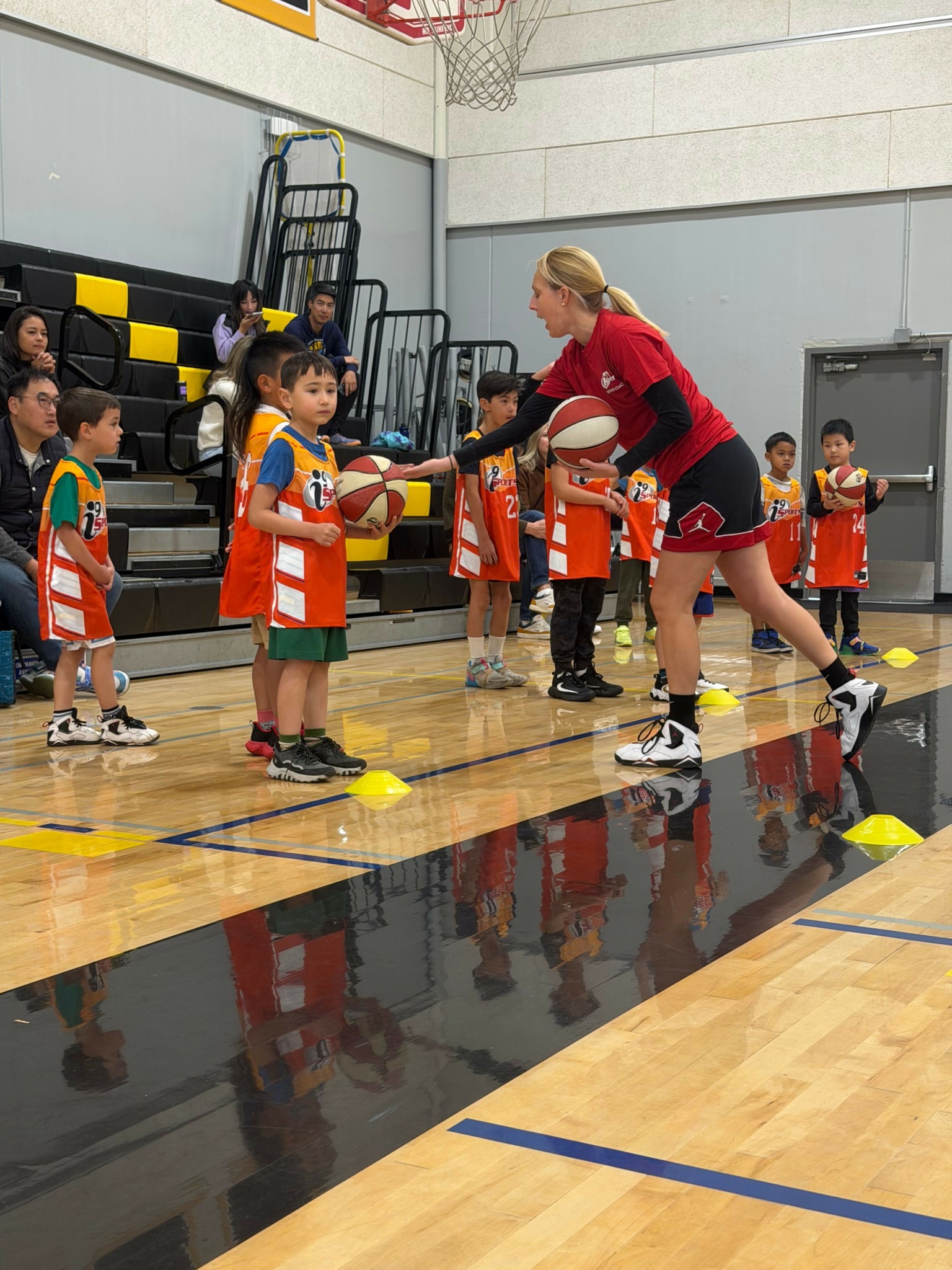 Children playing basketball in an indoor gym, some holding basketballs, with sports equipment and chairs in the background.