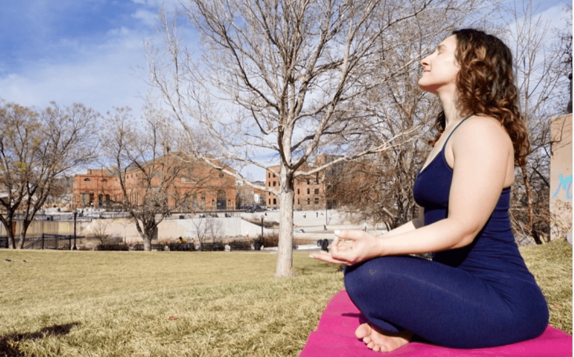 Woman practicing yoga outdoors on a pink mat, sitting cross-legged with eyes closed, trees and buildings in background.