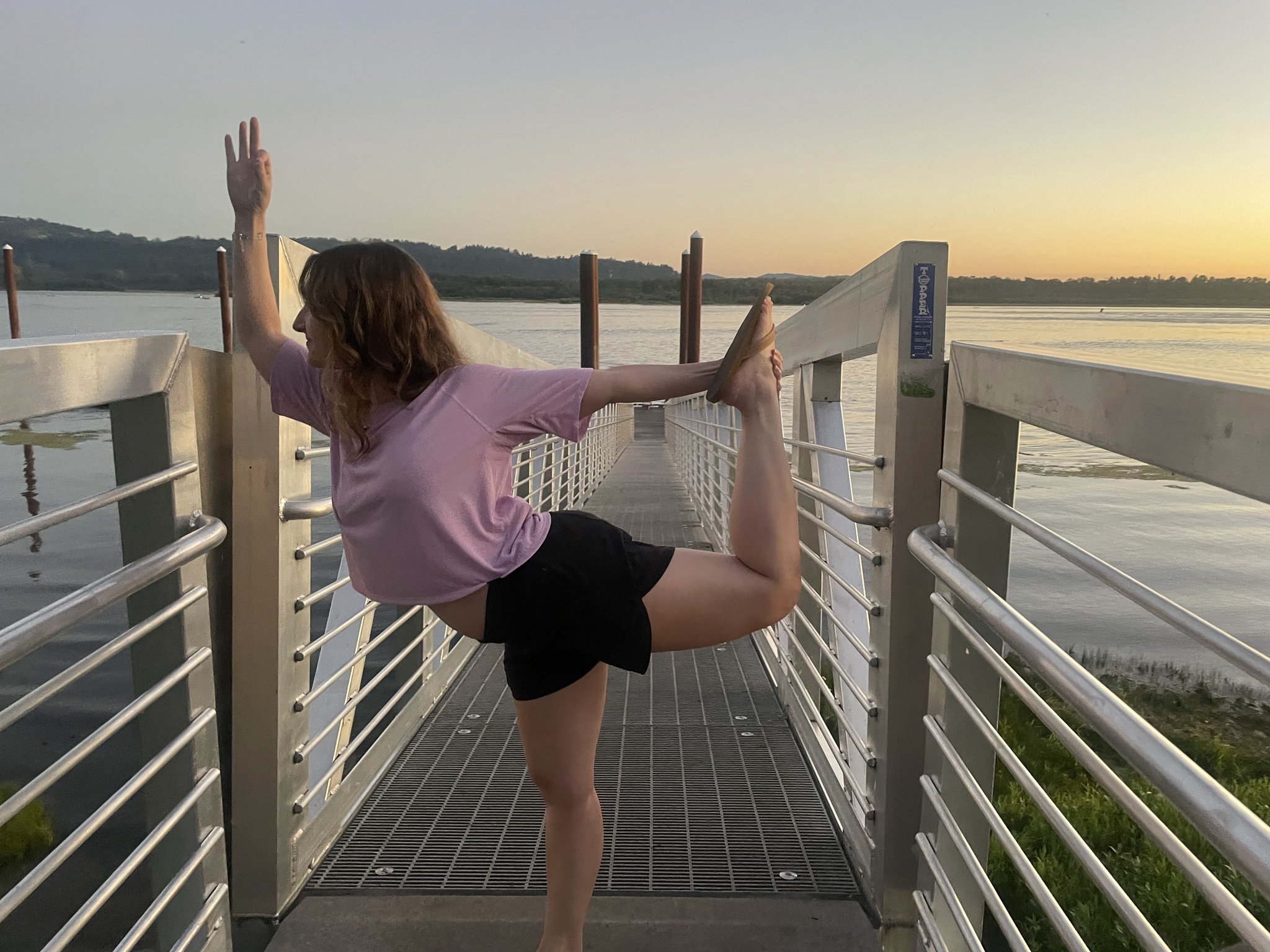 Person in pink top and black shorts doing a yoga pose on a dock by the water at sunset.