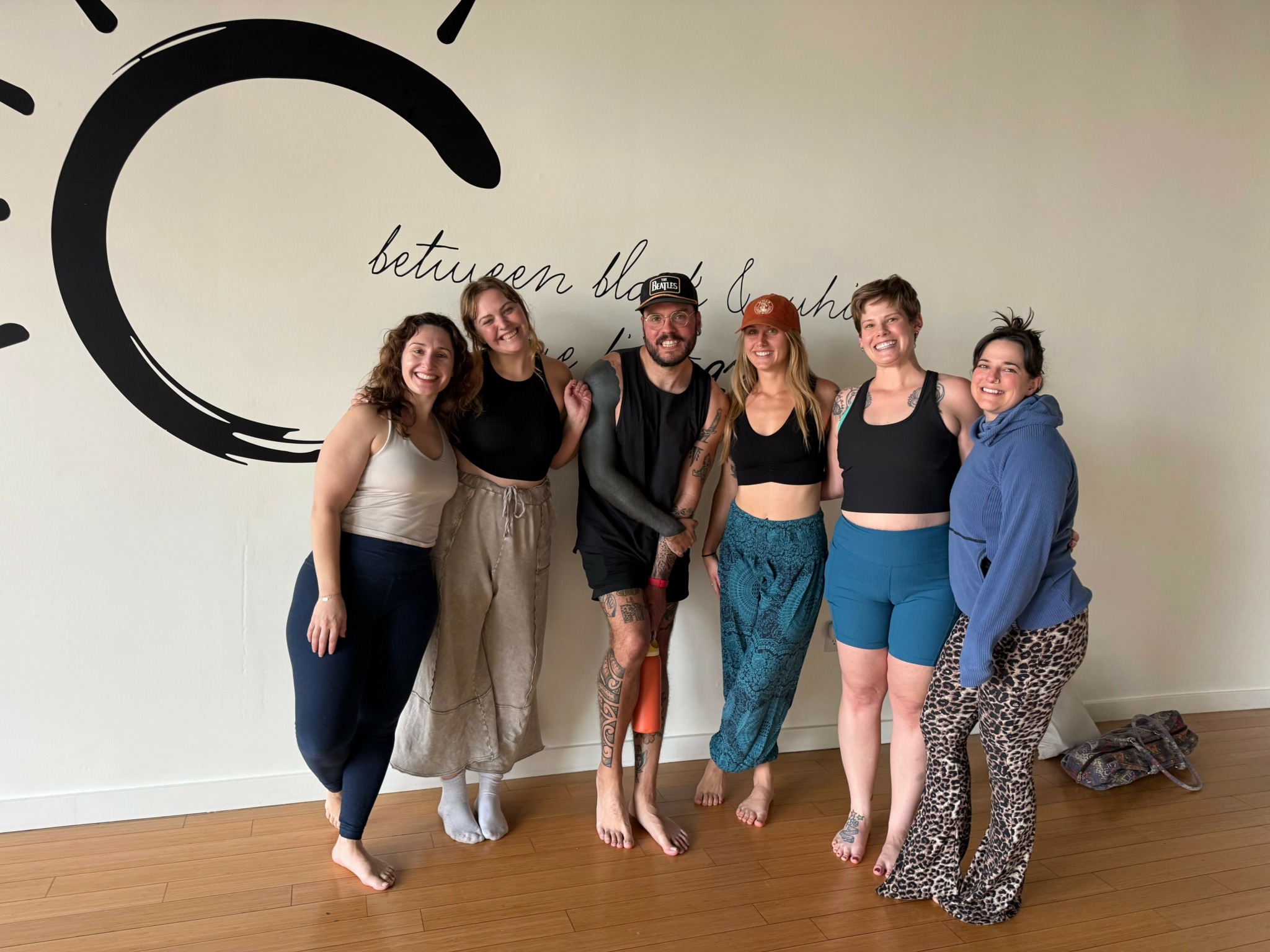 Six people standing together indoors, smiling, with a wall and wooden floor in the background.