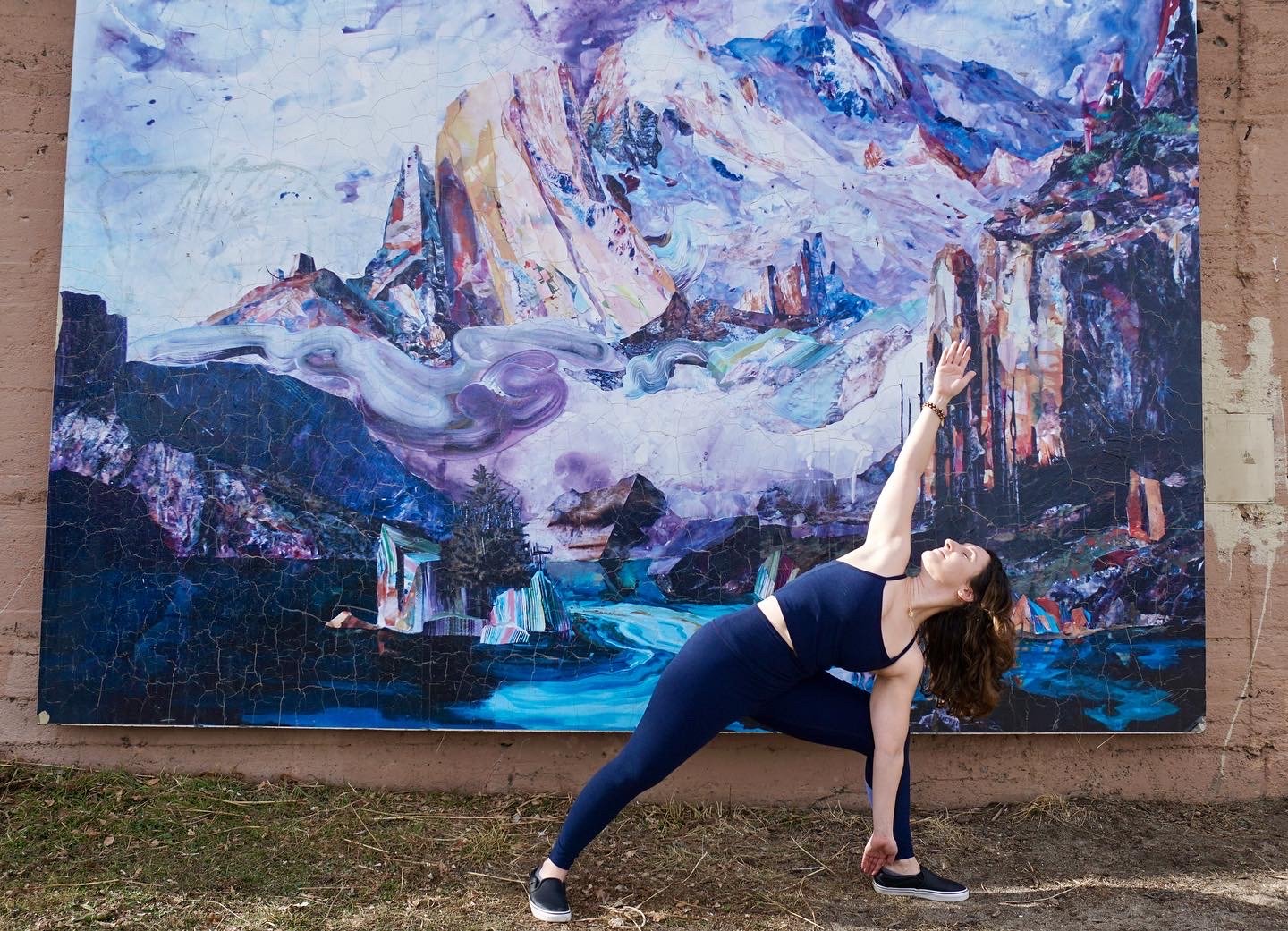 Woman performing yoga pose outdoors in front of a large colorful mountain landscape painting, with one arm extended upward and the other hand on the ground.