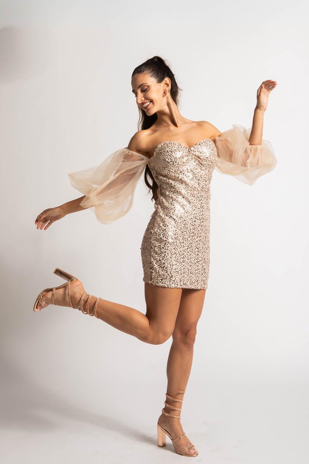 Woman in a sparkly dress and high heels poses with one leg raised and arms outstretched, smiling.