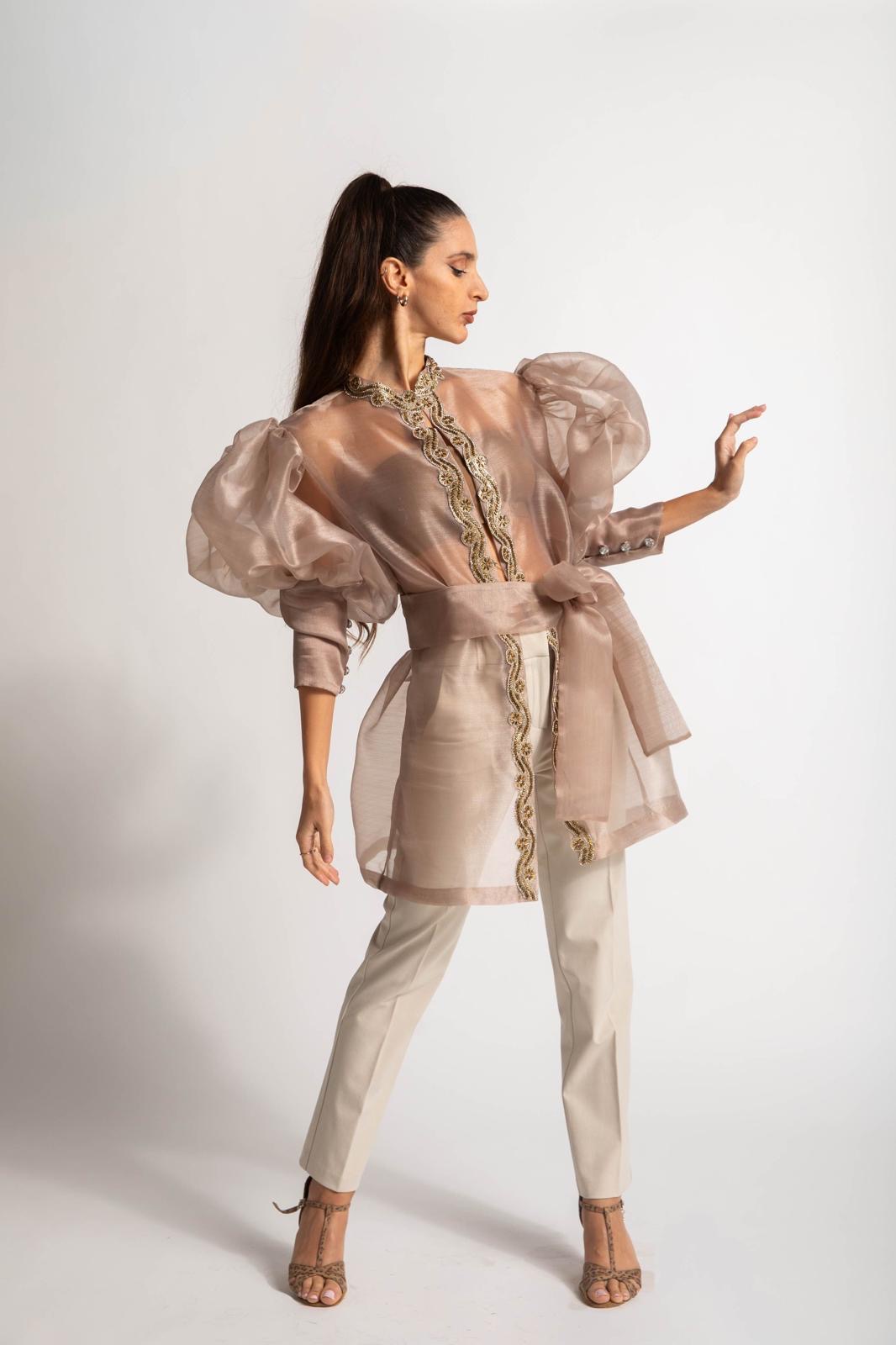 Woman in beige outfit with puffed sleeves and high heels, standing sideways with hand raised, against a plain background.