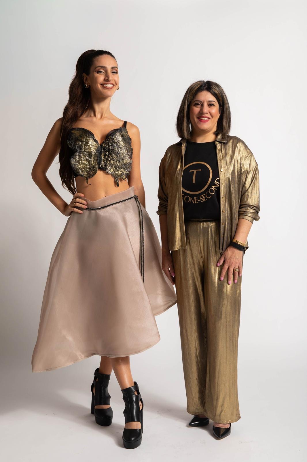 Two women standing together, smiling, against a plain white background. One wears a black and gold top with a beige skirt, the other a gold jacket and pants.