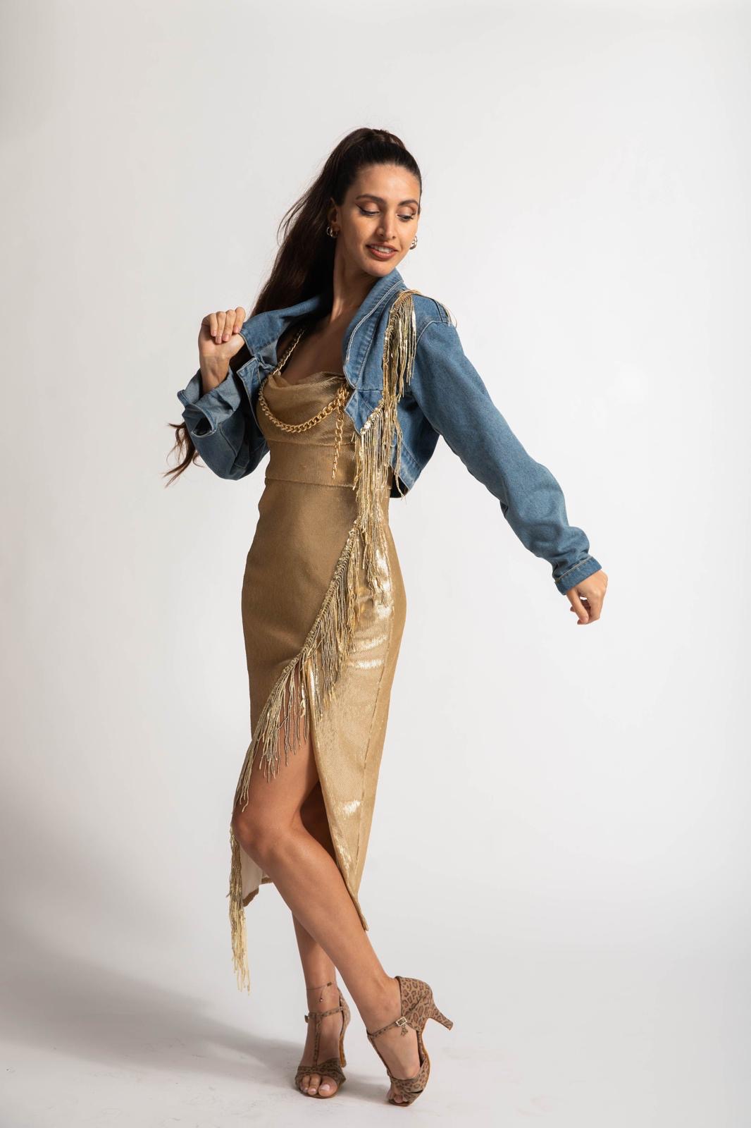 Woman in a gold dress and denim jacket poses with eyes closed, one hand on her shoulder, against a plain white background.