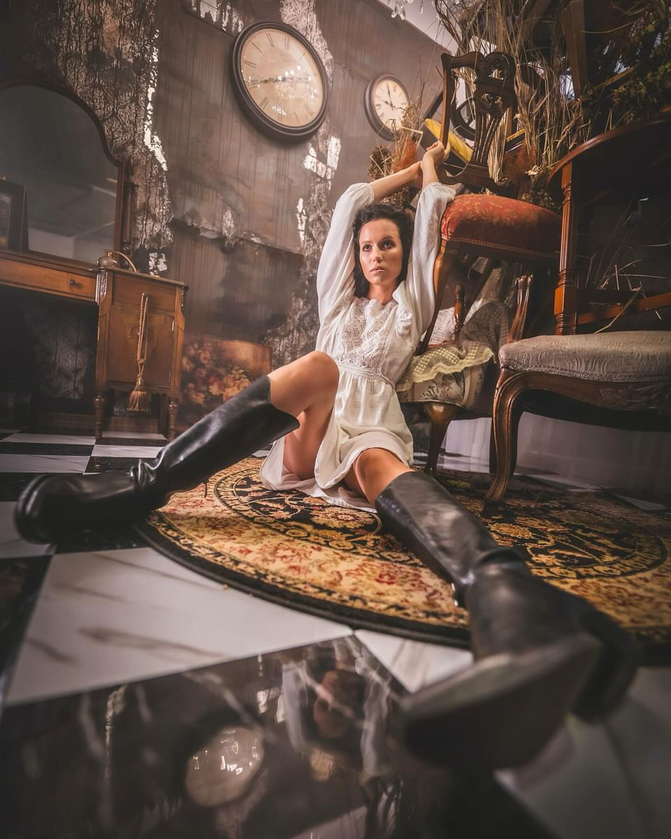 Woman in a white dress and black boots sitting on a rug, reaching up with her arms in a vintage room.
