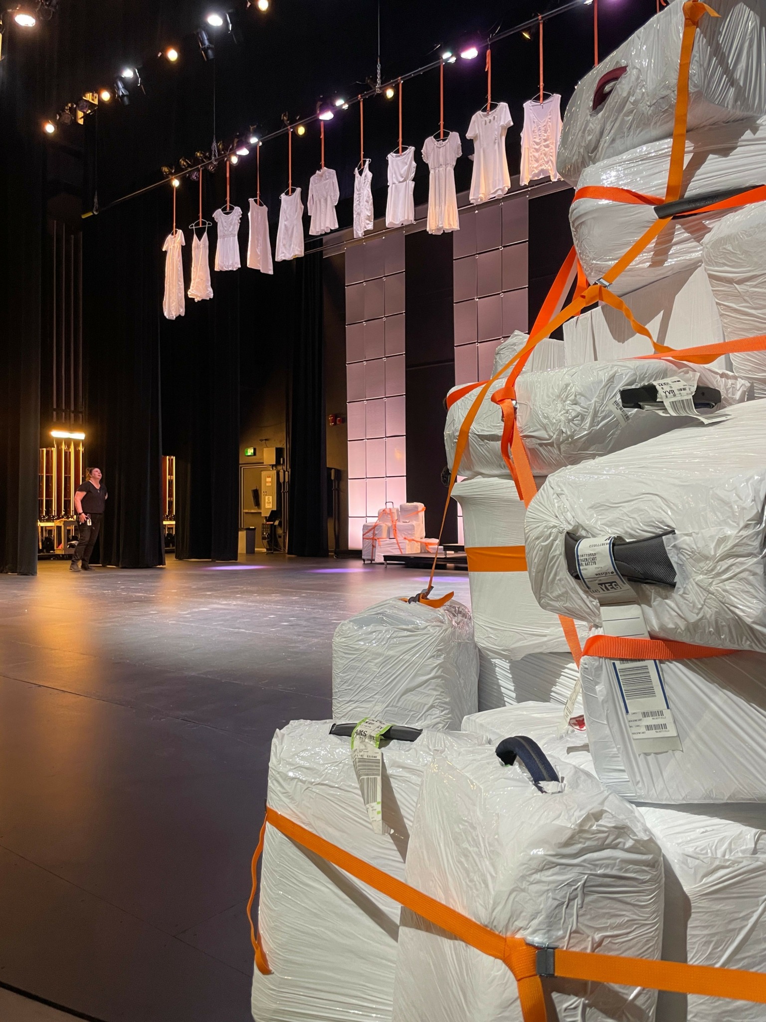 Large packages wrapped in white material with orange straps are stacked in a room with a polished floor and string lights.