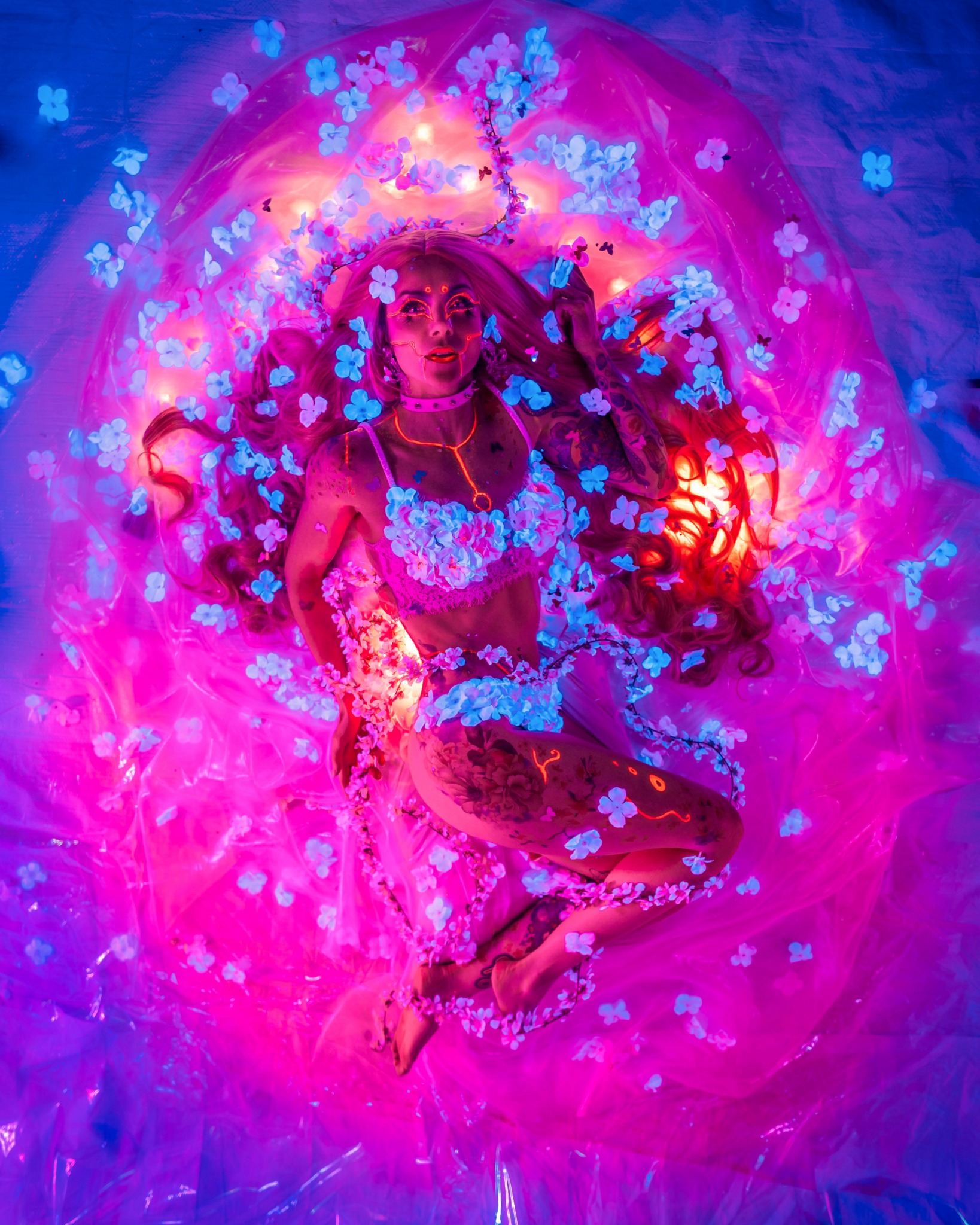 Person lying in a colorful, glowing environment with pink, purple, and blue lights, surrounded by floating flower-like shapes.