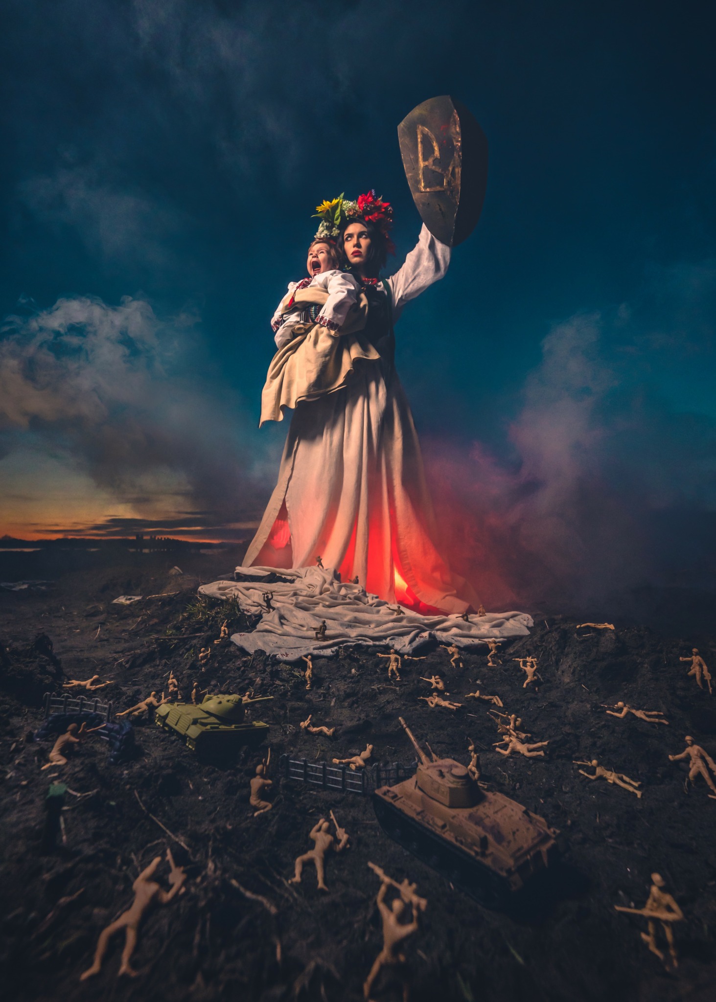 Giant woman in traditional dress holding a shield and flowers, standing on a rocky landscape with a dark sky, illuminated from below.