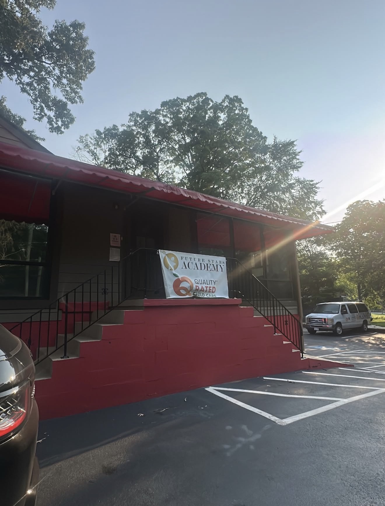 Building with red stairs and a banner, parking lot with cars, trees in background, sunlight shining from right side.