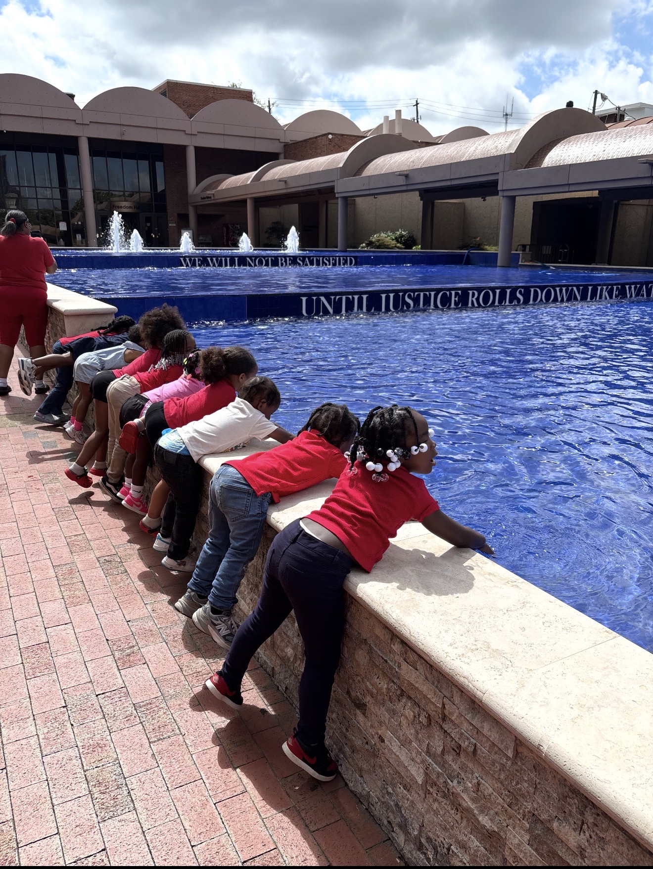 Children leaning over a fountain's edge, looking into the water, with a building and cloudy sky in background.