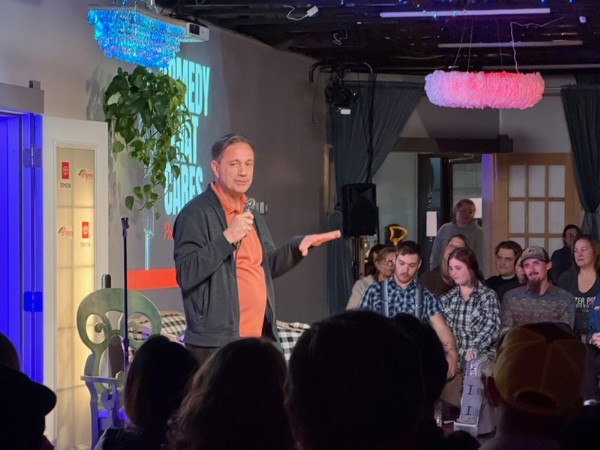 Man speaking on stage with audience seated, indoor setting, colorful lighting, plants, and decorations.
