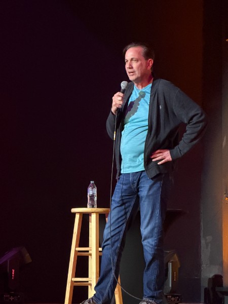 Man standing on stage holding a microphone, with a water bottle on a stool beside him, dark background.