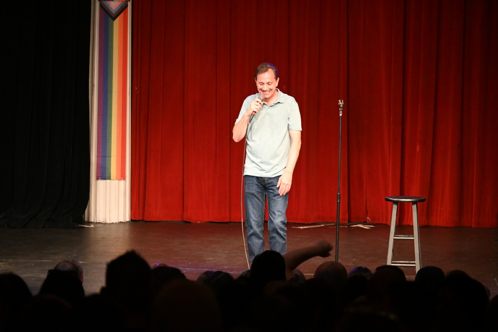 Man standing on stage with red curtain, holding microphone, smiling, with a stool nearby, audience in foreground.