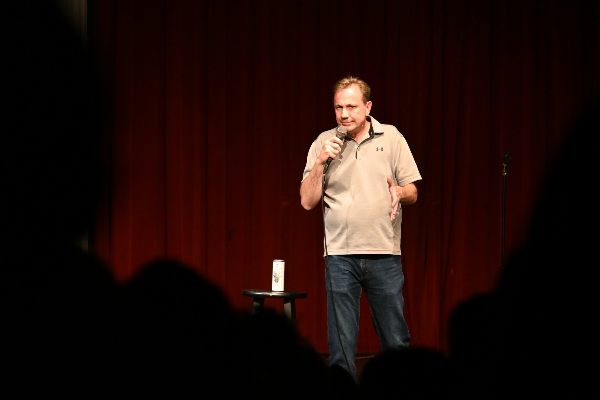 Man standing on stage with a microphone, red curtain background, audience silhouettes in foreground.