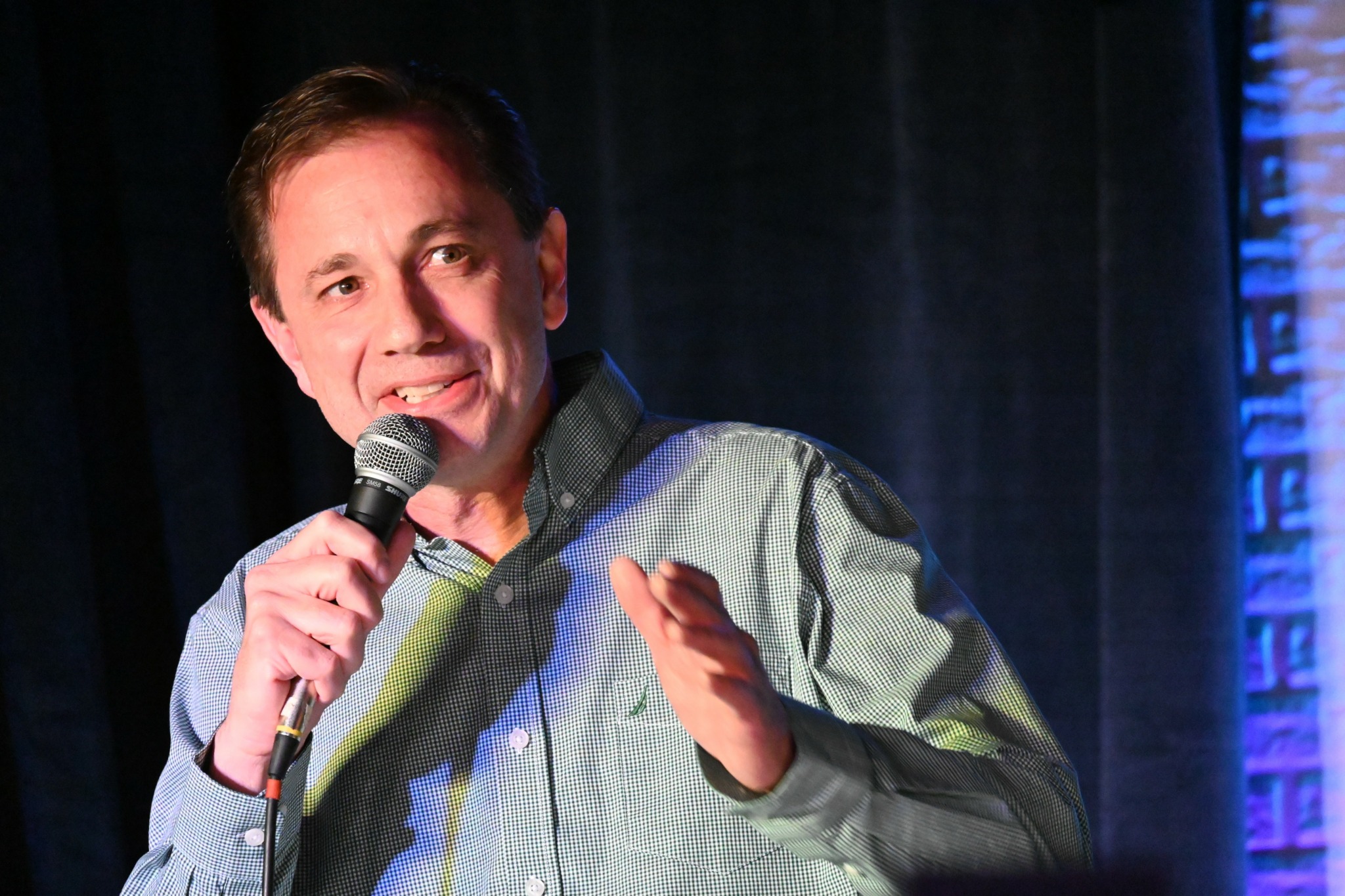 Man holding a microphone, speaking on stage with a dark background and blue lighting, gesturing with his hand.
