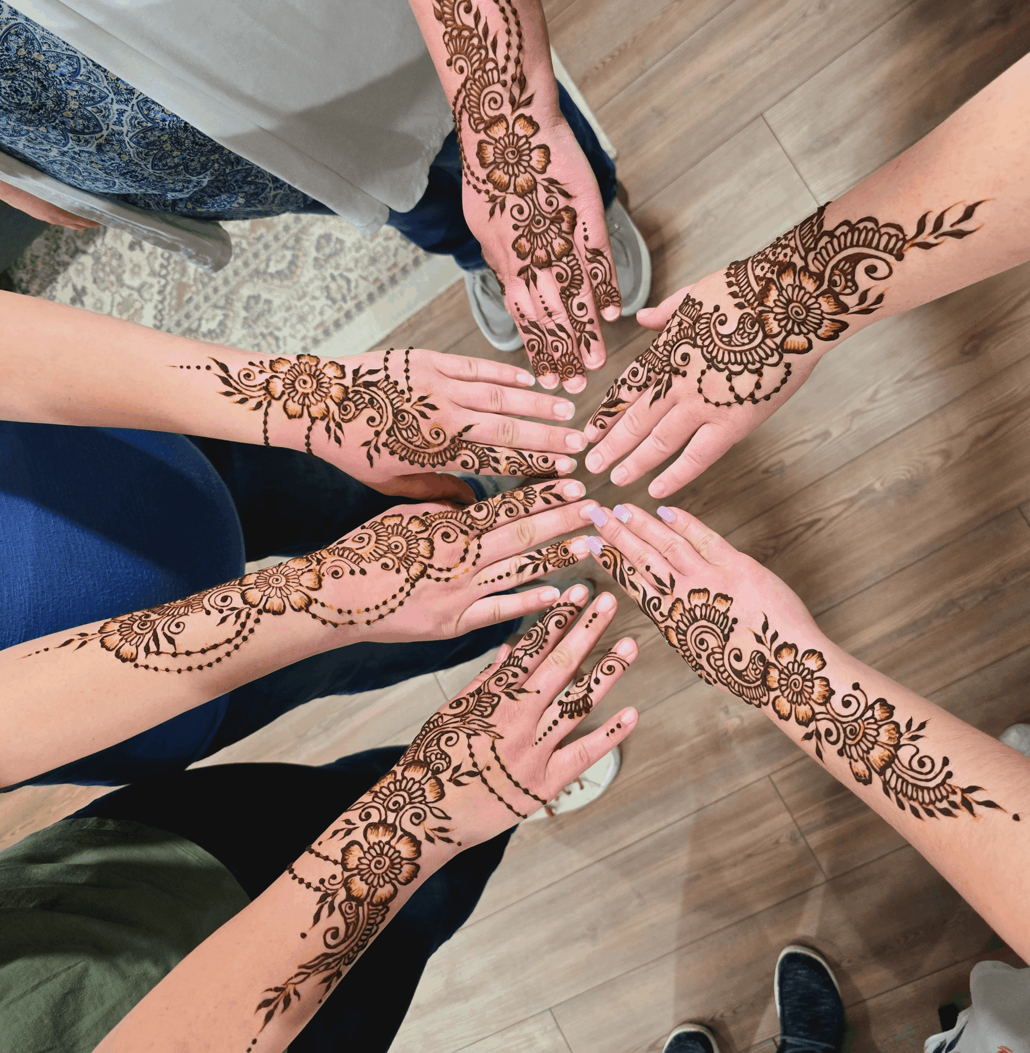 Six hands with intricate henna designs extended towards each other in a circle on a wooden floor.