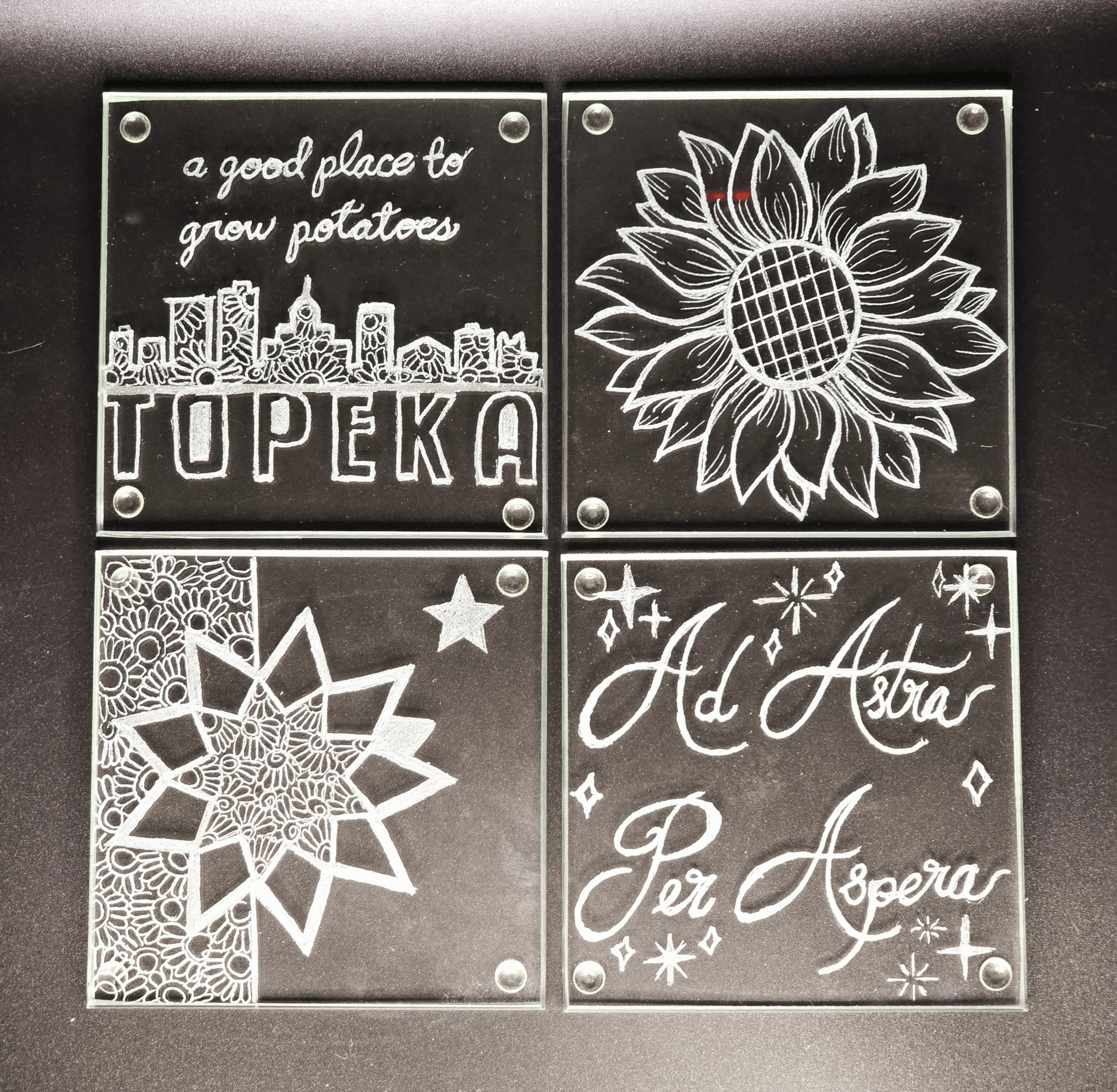 Four glass plaques with white drawings and text, including a cityscape, sunflower, star, and a quote in Italian, on a black background.