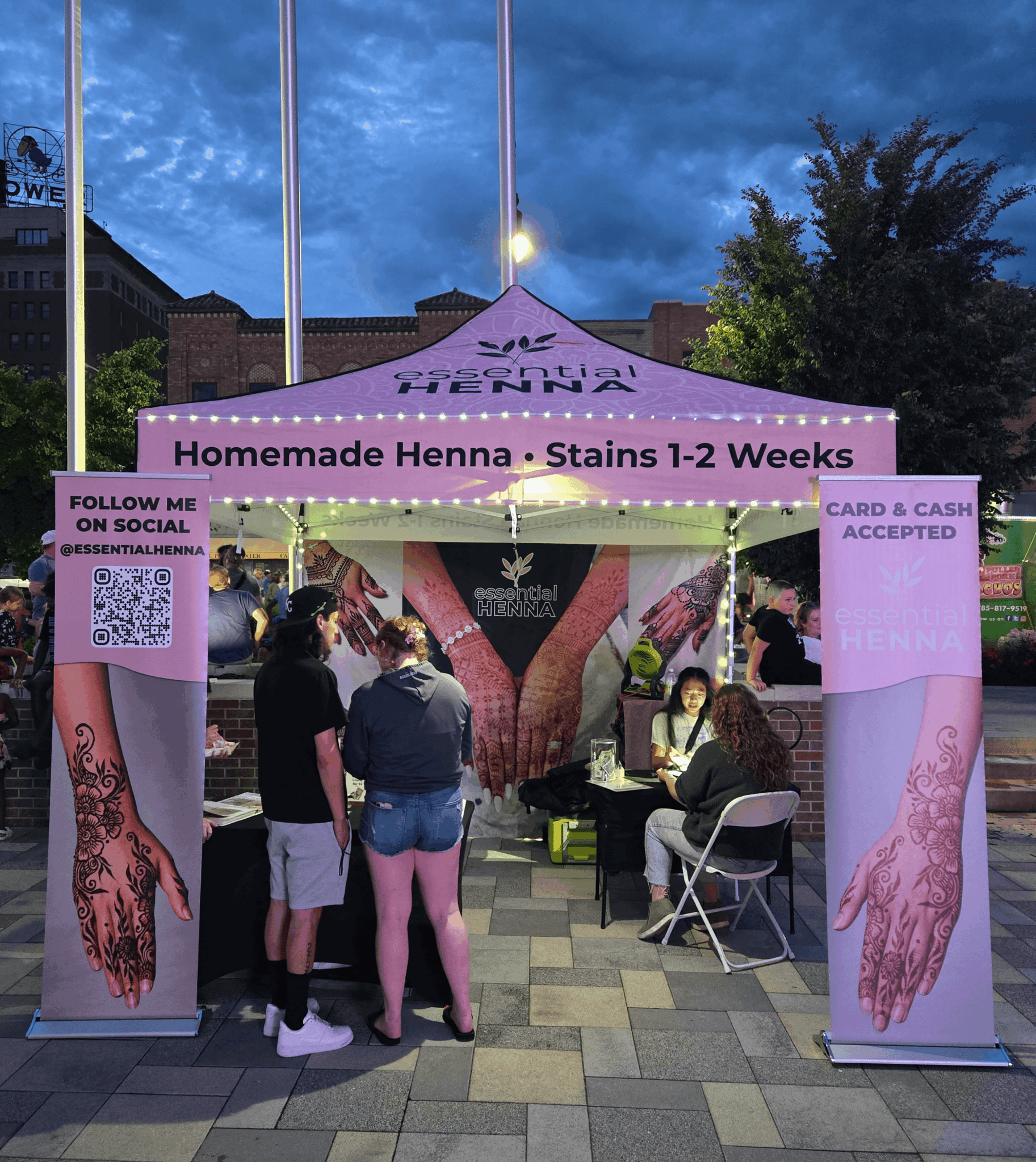 Pink tent selling homemade henna with people gathered around, city buildings in background, evening sky, and signs about henna and payment.