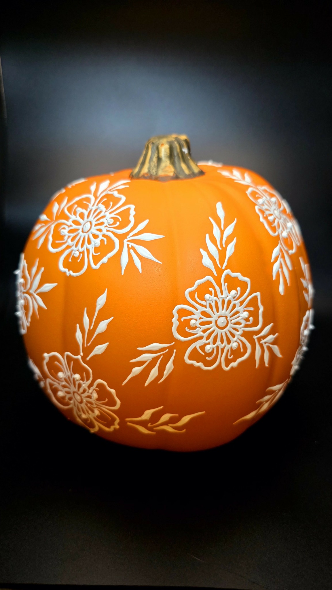 Orange pumpkin with white floral patterns, gold stem, on a dark background.