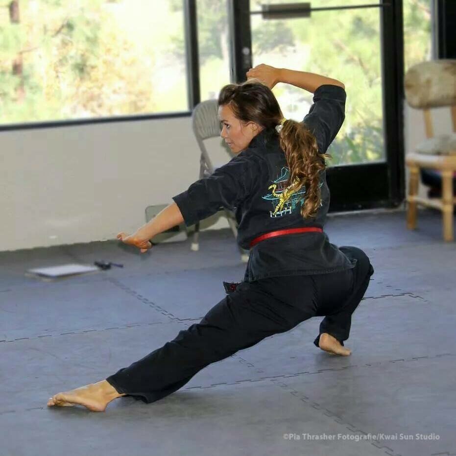 Woman practicing martial arts in a room with large windows, wearing a black uniform, in a low stance with one arm extended.