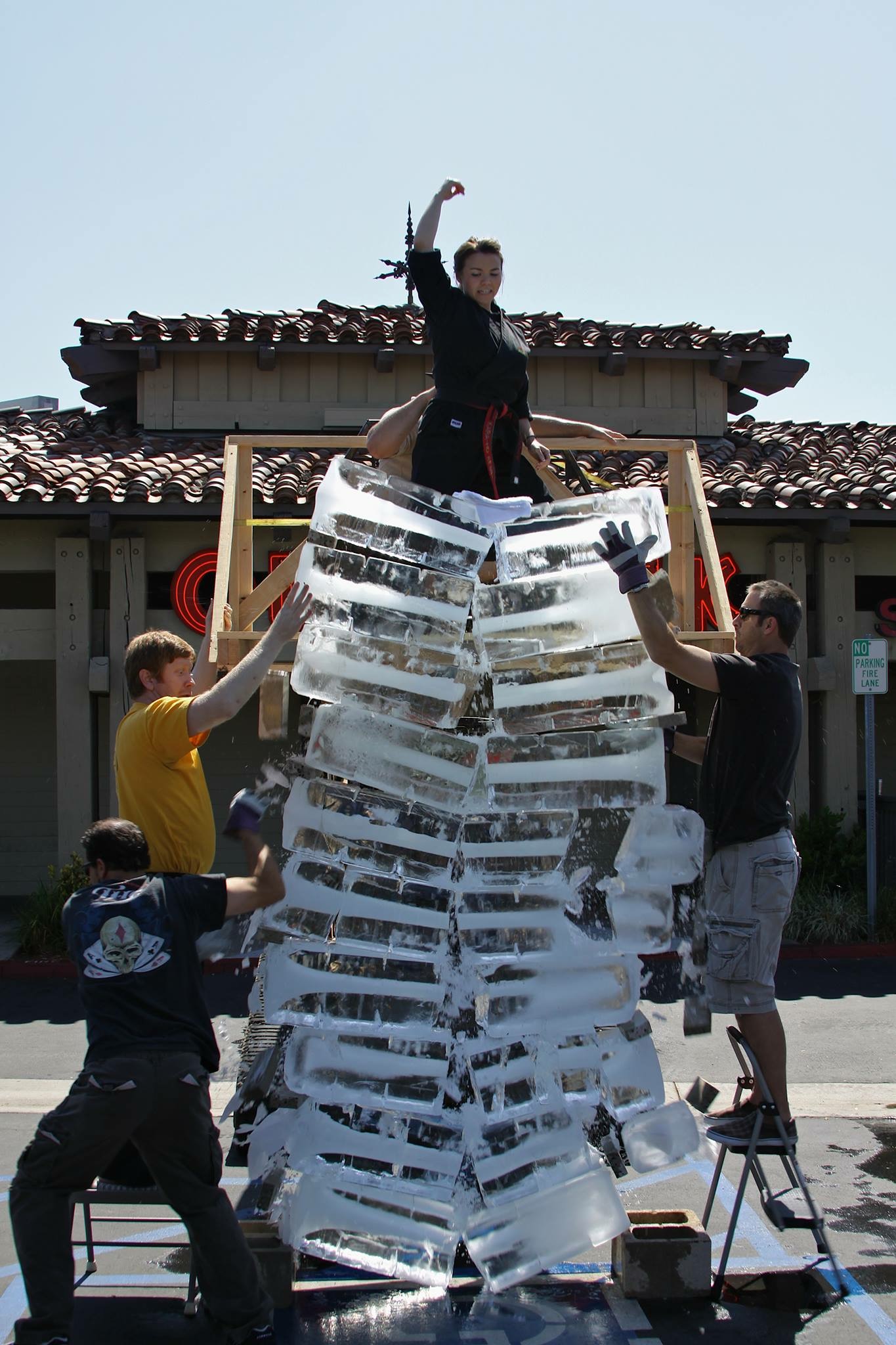 Person standing on a tall structure made of stacked ice blocks, with three people supporting it, outdoors under a clear sky.