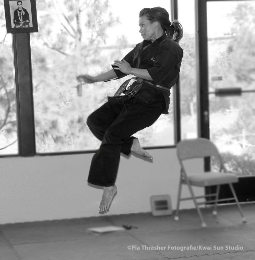 Woman practicing martial arts in a dojo, mid-air with a focused expression, wearing a black uniform, near a window and chair.