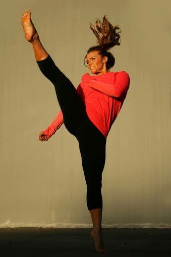 Dancer in red top and black pants performing a high kick with hair flying, against a plain background.
