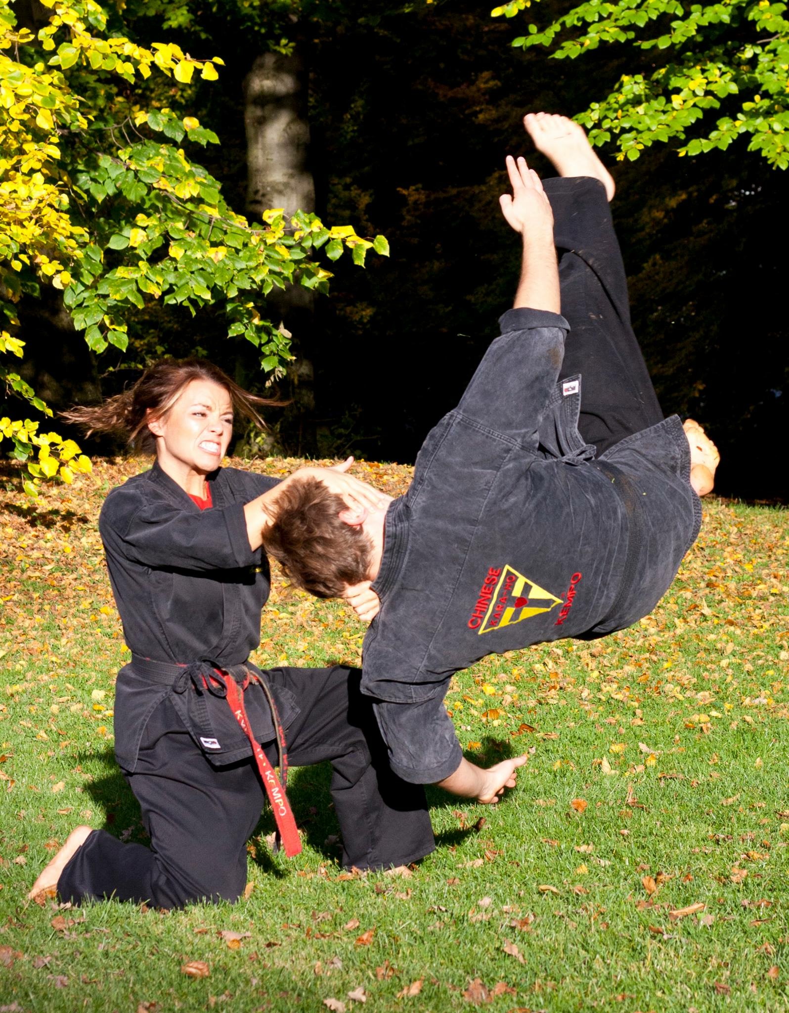 Two people practicing martial arts outdoors on grass, one person in a black uniform executing a flip, the other kneeling.