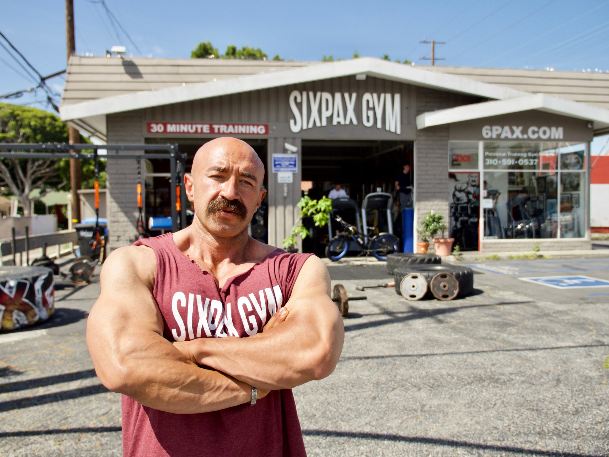 Man with muscular arms and a mustache stands outside SixPax Gym in sunlight, crossing arms, in front of gym entrance.