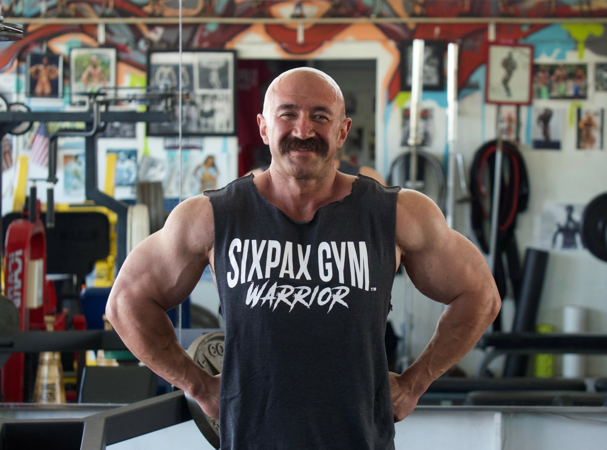 Smiling man with muscular arms in gym, wearing a sleeveless shirt, standing with hands on hips, gym equipment in background.