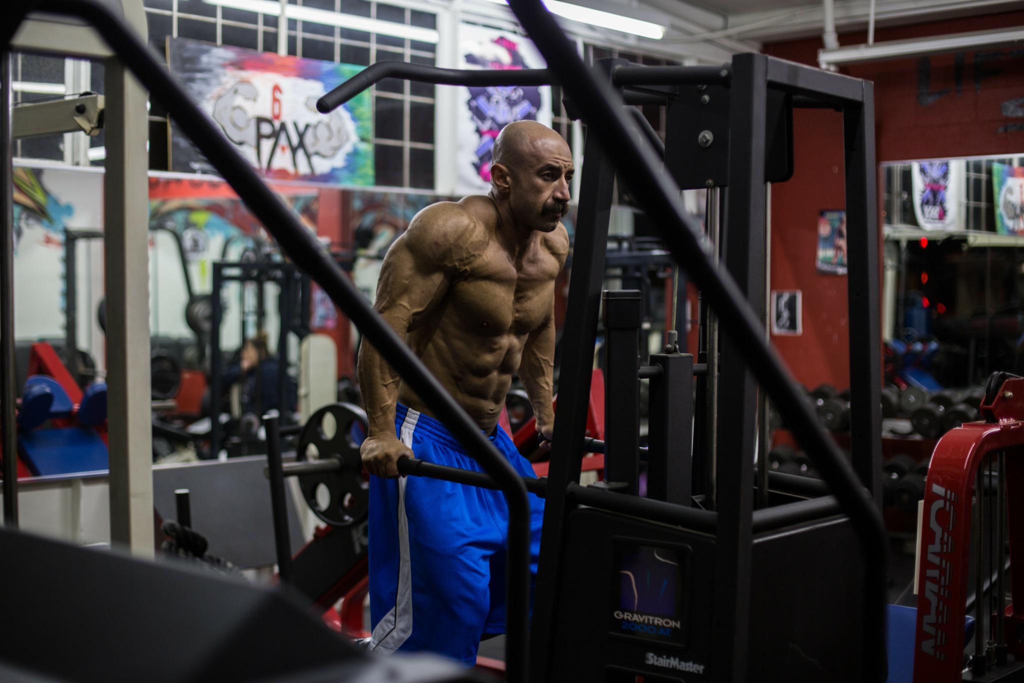 A muscular man with a shaved head and beard exercises in a gym, lifting weights with graffiti on the walls behind him.