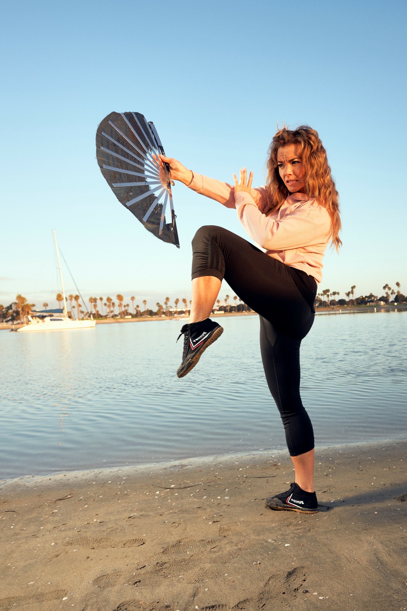 Woman standing on beach holding a fan, lifting her leg, with water and boats in background.