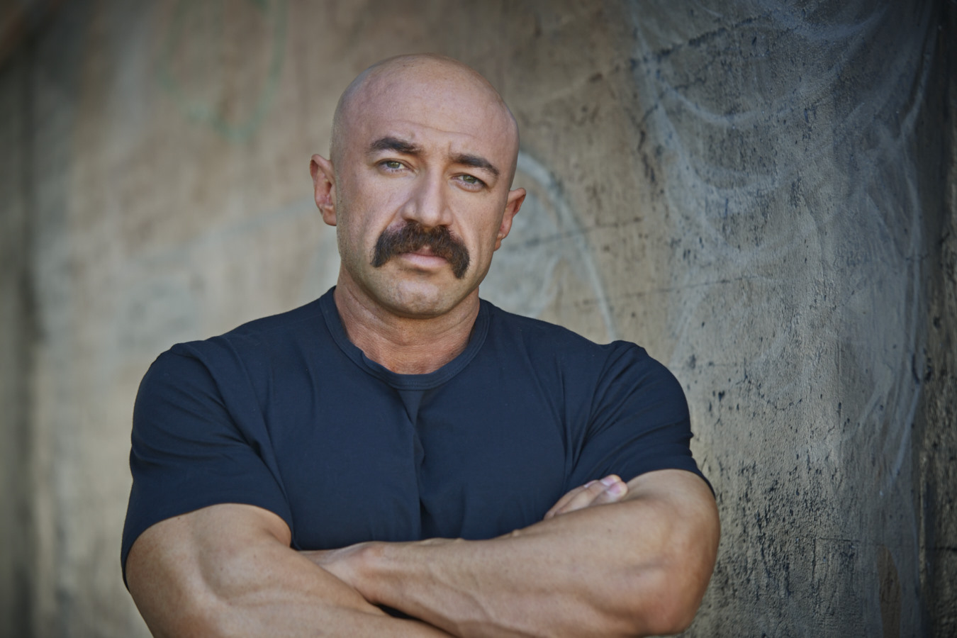 Man with a mustache and bald head, wearing a dark blue shirt, standing with arms crossed against a textured wall.