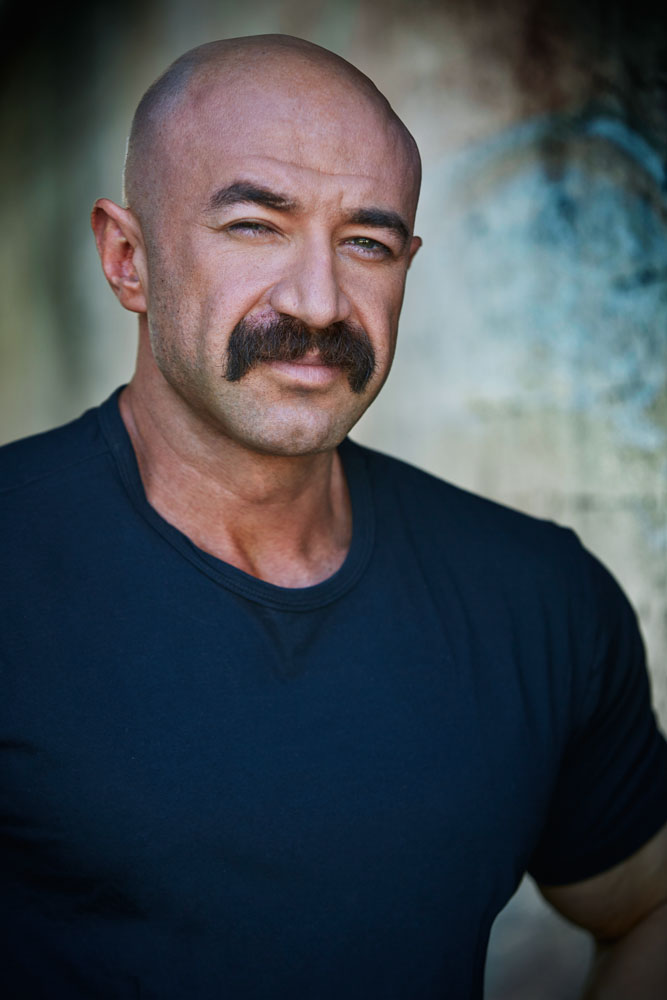 Man with a bald head and mustache wearing a dark shirt, outdoors with blurred background