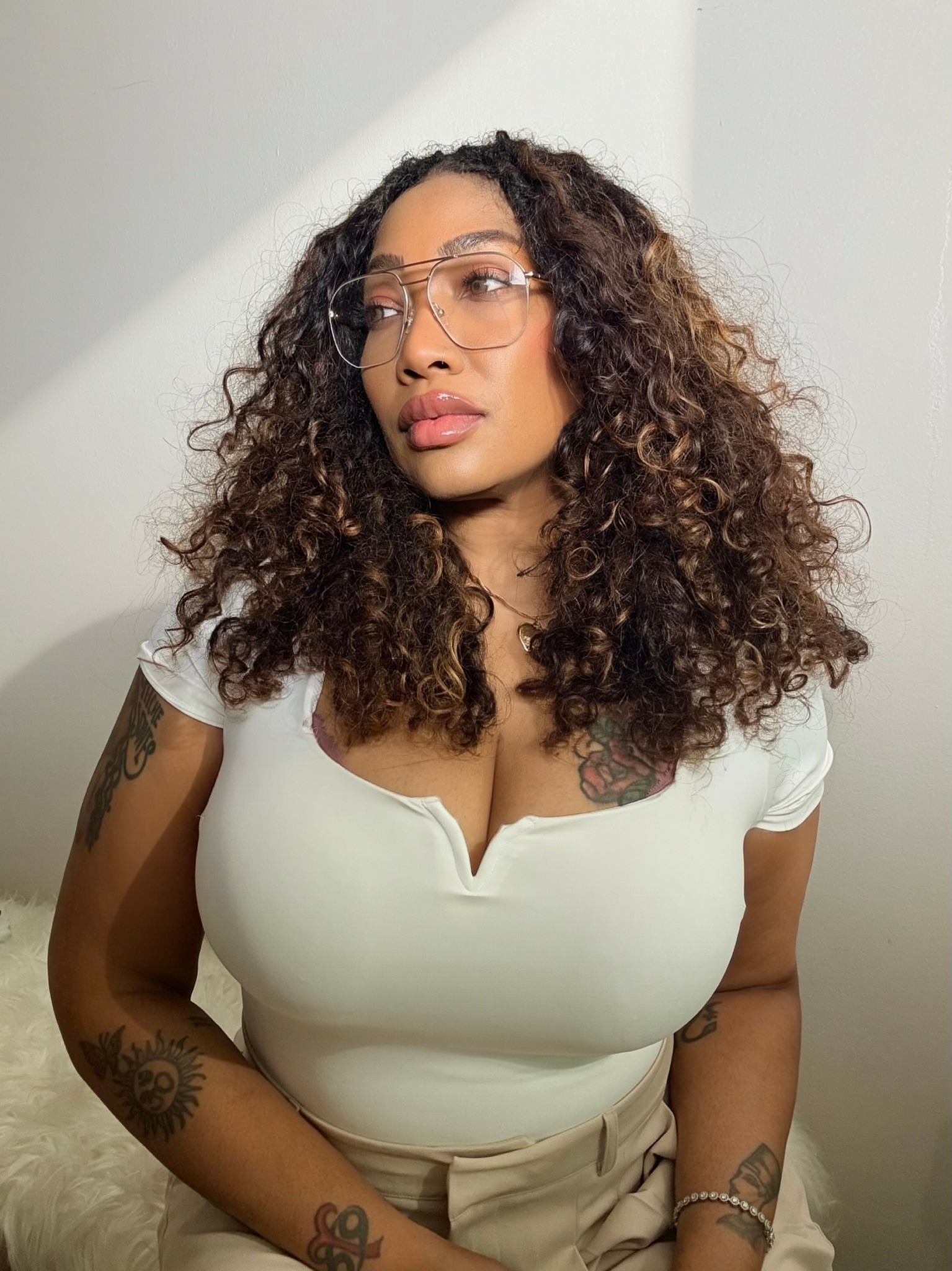 Woman with curly hair wearing glasses and a white top, looking to the side, against a plain background.