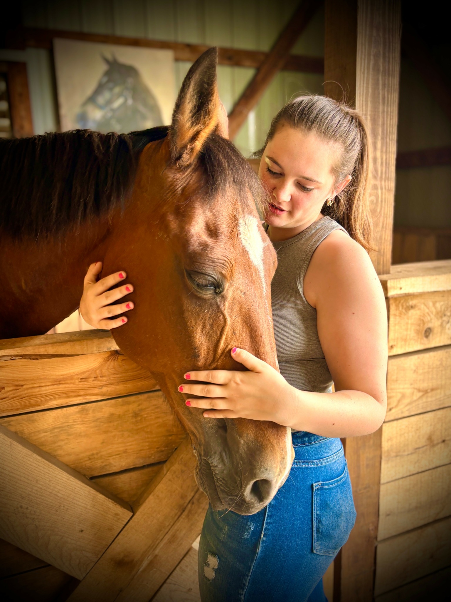 Young woman gently touches a horse's face inside a stable, wooden walls in background.