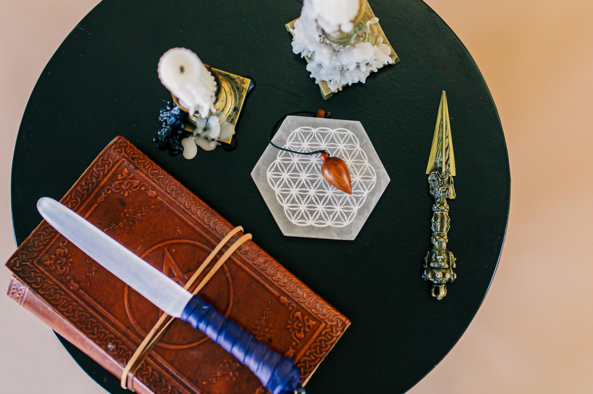 Objects on a round black table include a brown leather-bound book, a white feather, a shell, a hexagonal dish with a small object, and a decorative chain.