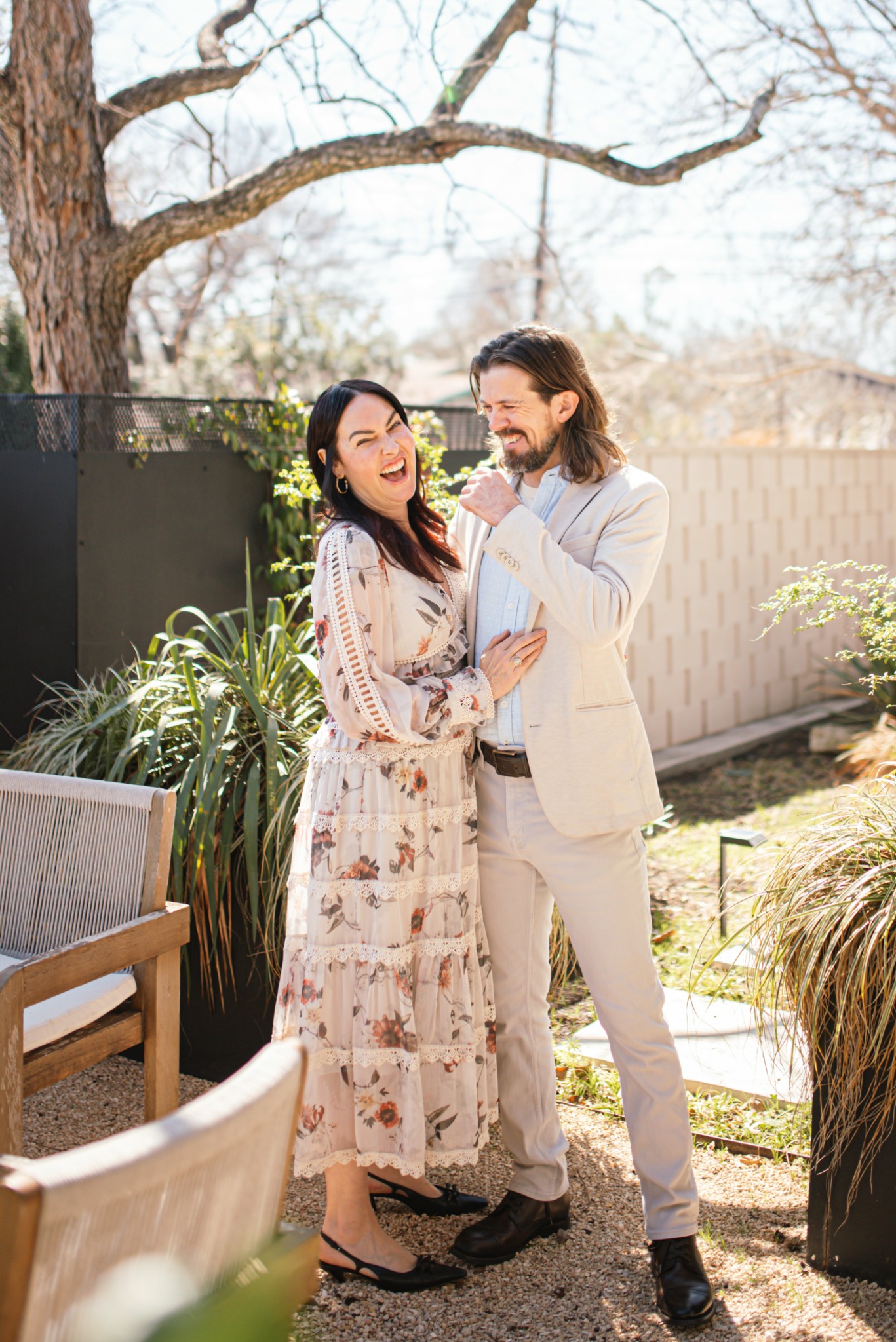 Two people smiling and talking outdoors in a garden, with trees and a fence in the background.