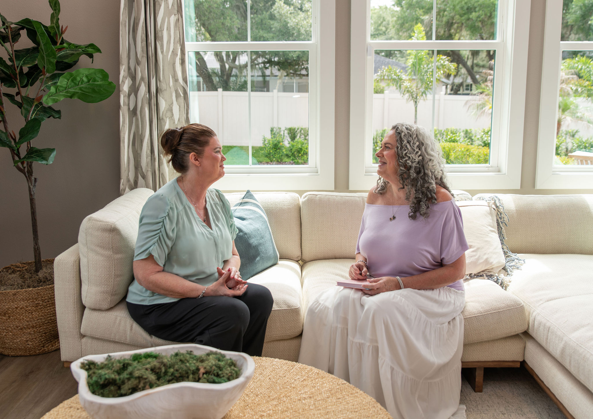 Two women sit on a white sectional sofa in a bright room with large windows, engaged in conversation.