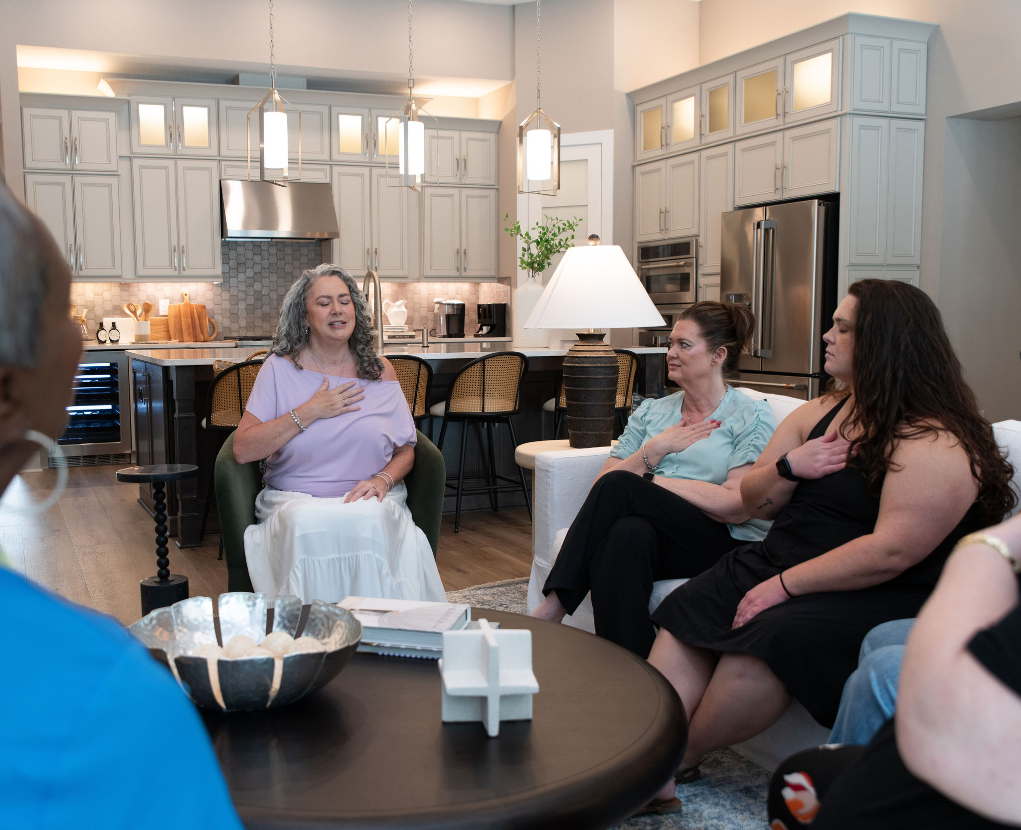 Group of women sitting and talking in a modern kitchen and living room area, with a woman speaking and others listening.