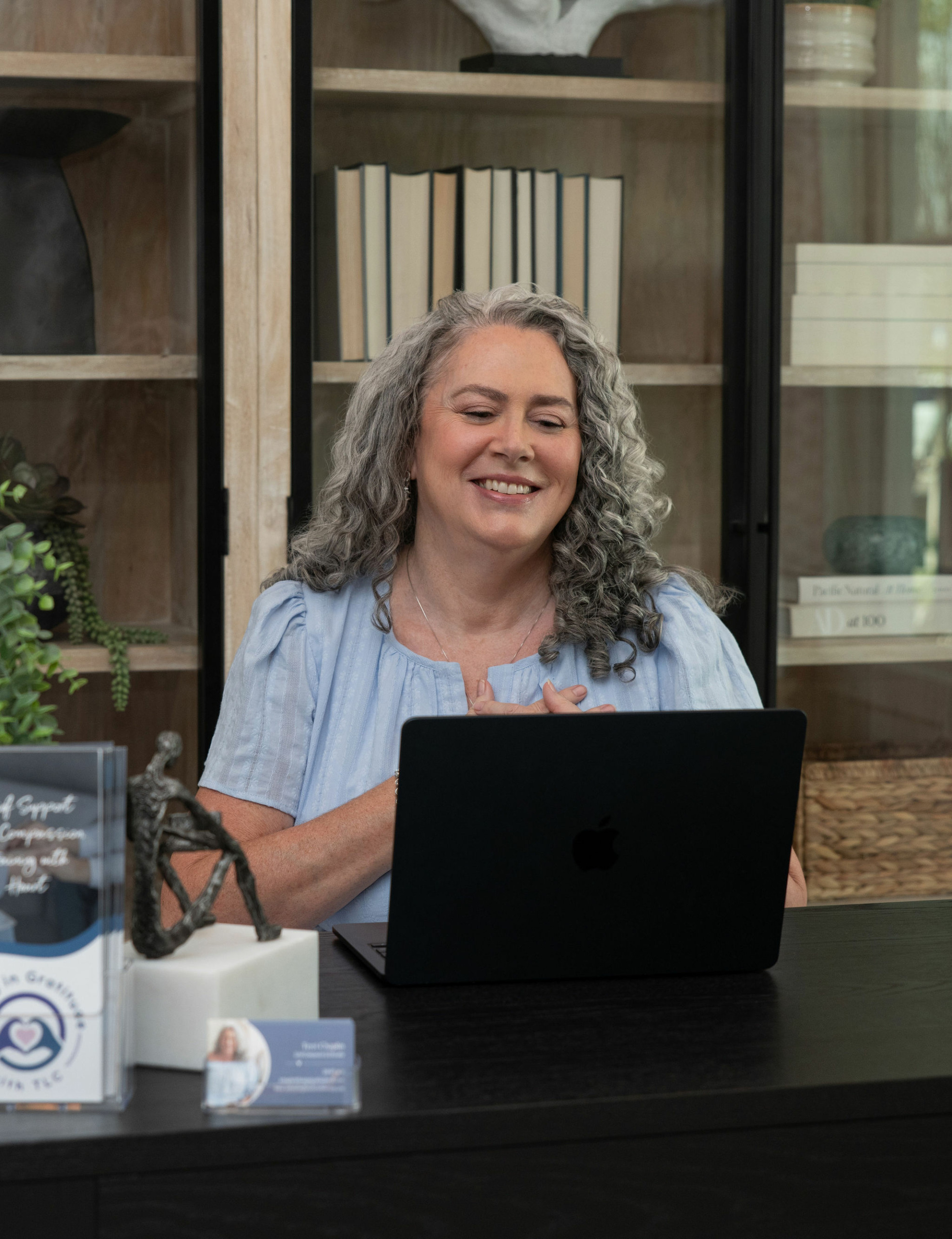 Smiling woman with curly gray hair sitting at a desk with a laptop, bookshelves in the background.