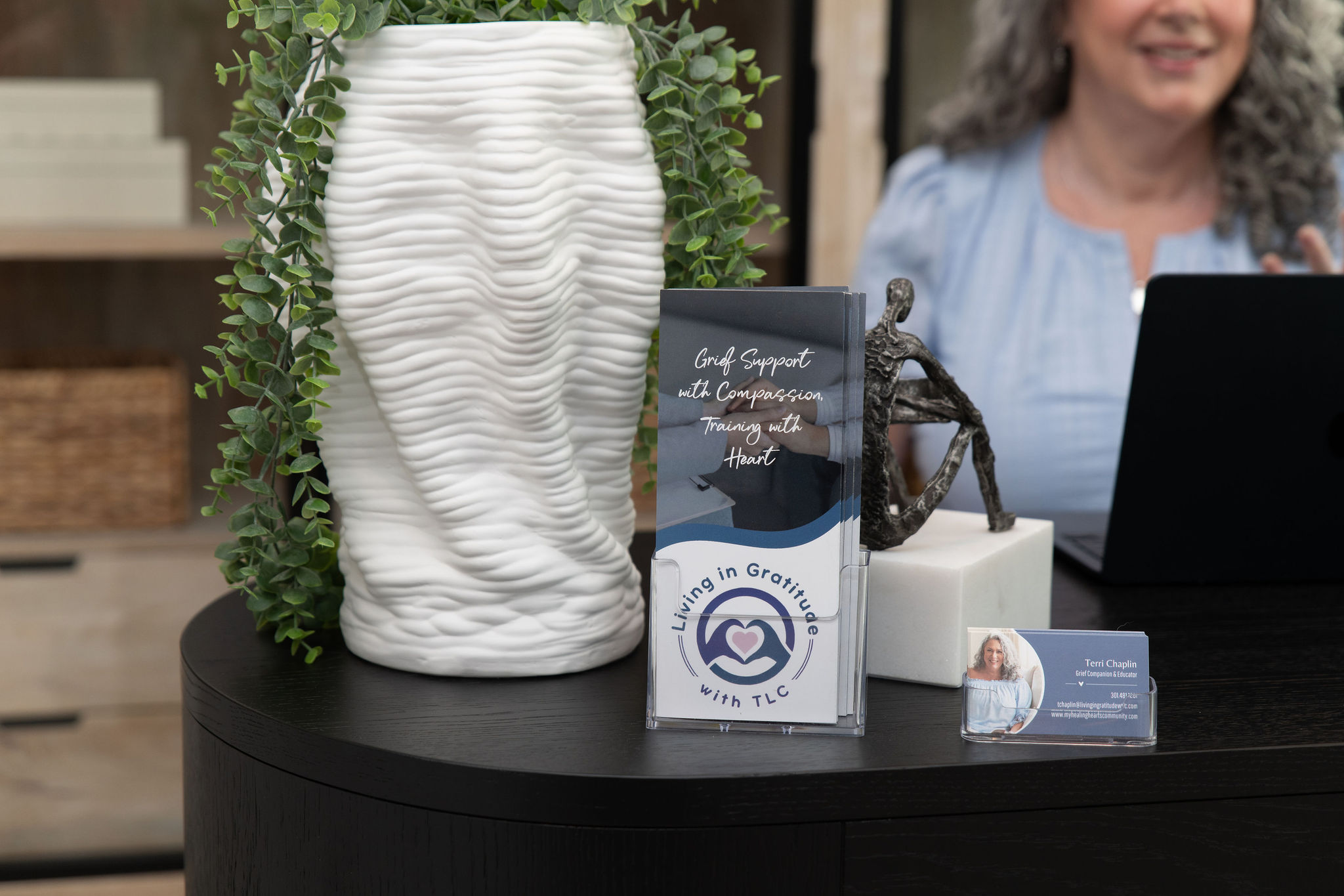 Person smiling at a desk with a large textured white vase, a mug, and business cards on a black table.