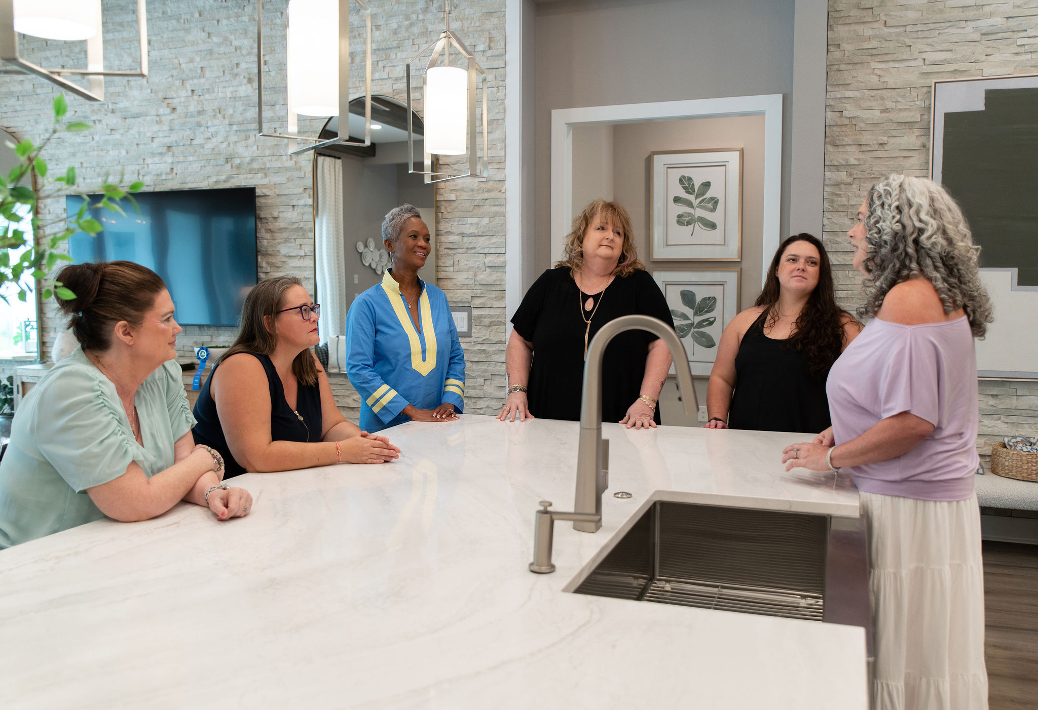 Five women standing and sitting around a kitchen island in a modern kitchen, engaged in conversation.