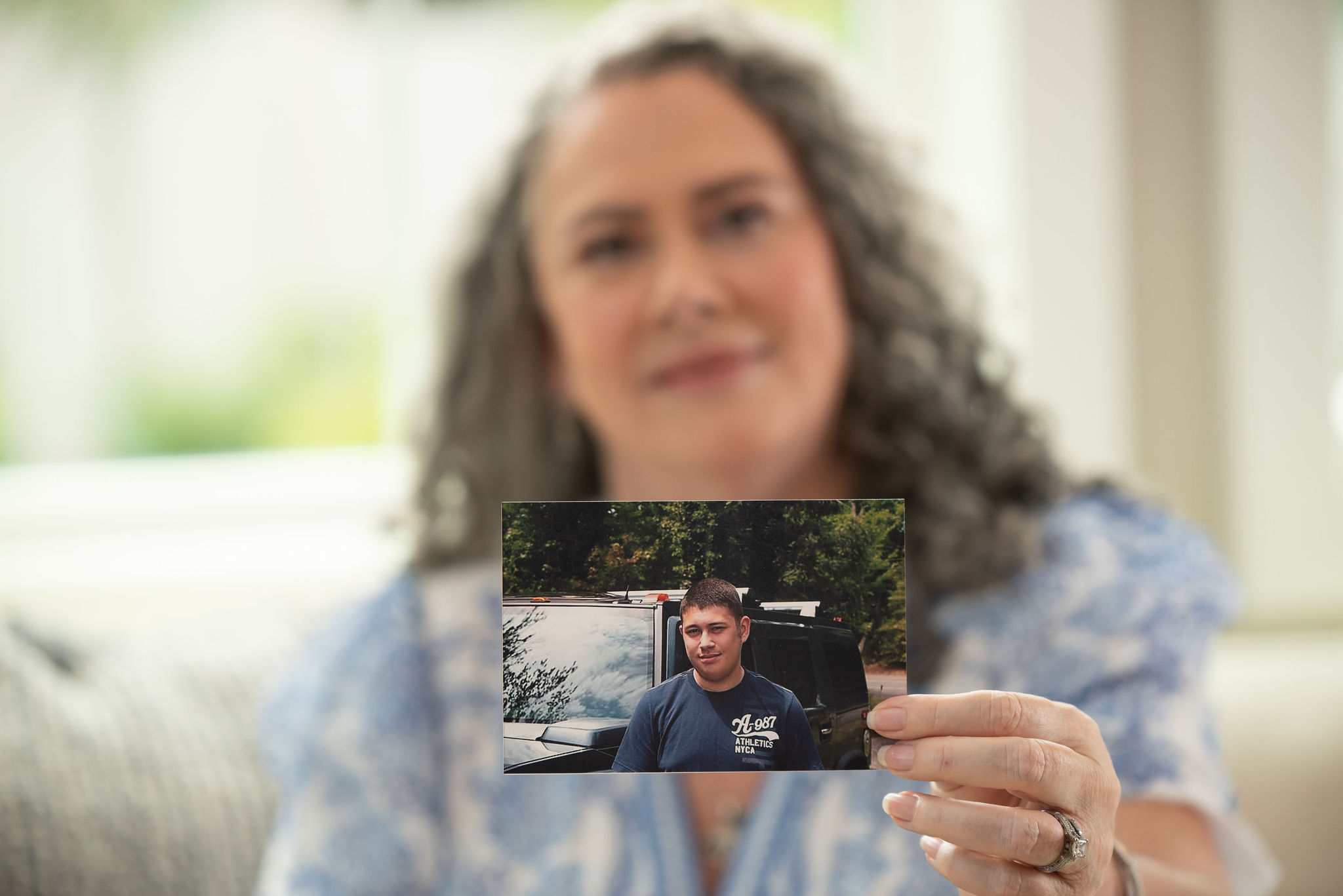 A woman with curly hair holds a small photo of a man outdoors near water, smiling softly. Background is blurred.