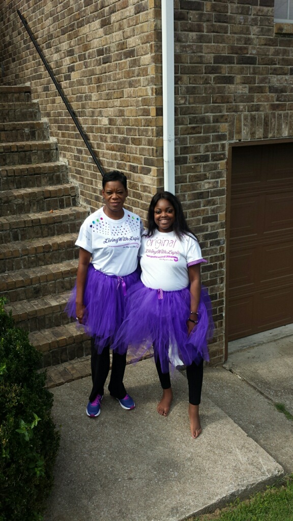 Two women stand outside near a brick wall, wearing purple tutus and white T-shirts, smiling at the camera.