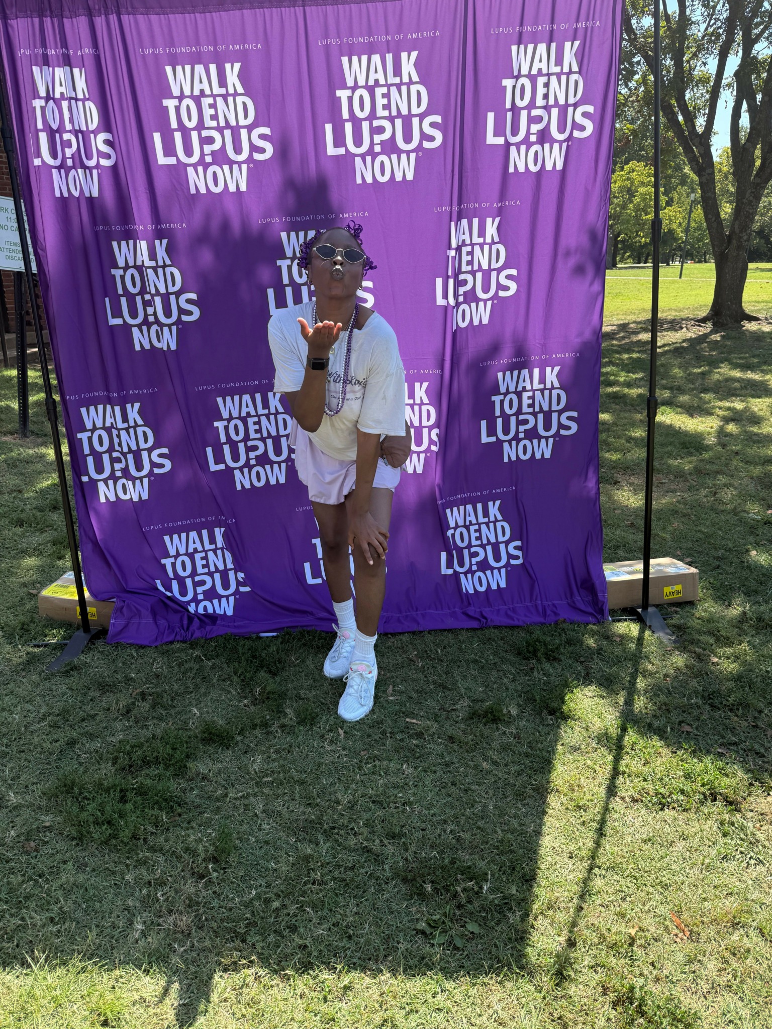 Person standing in front of a purple banner with white text in a park, trees and grass visible.