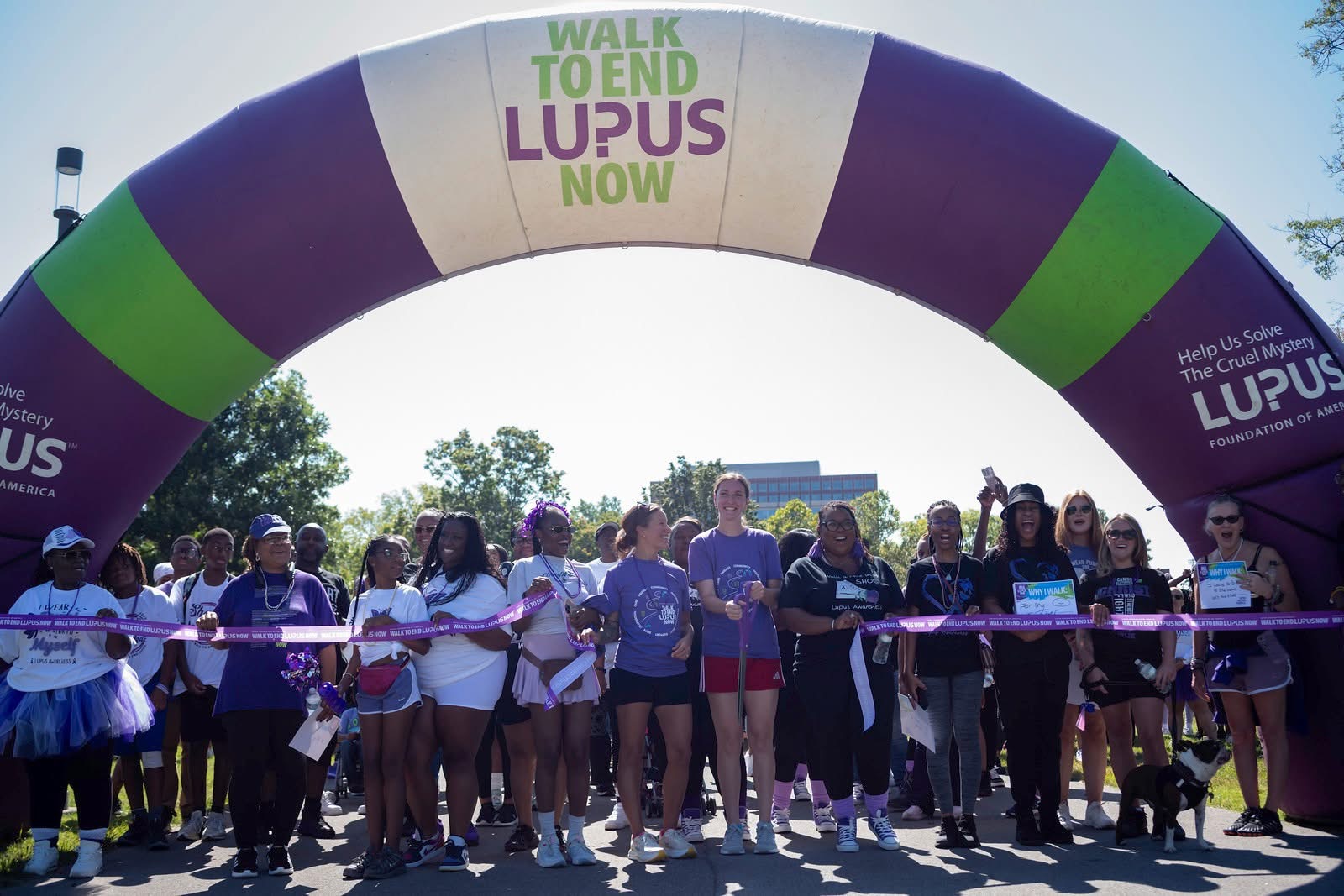 Group of people participating in a walk event under an arch with a sign promoting lupus awareness, outdoors on a sunny day.